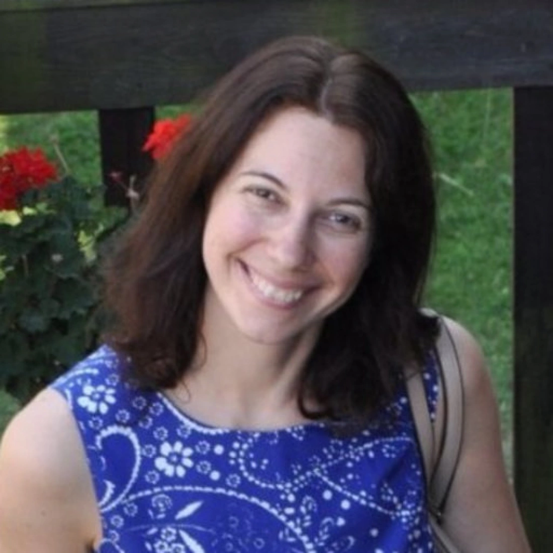 Kobieta w niebieskiej bluzce uśmiecha się, siedząc przed drewnianym ogrodzeniem w tle z kwiatami.