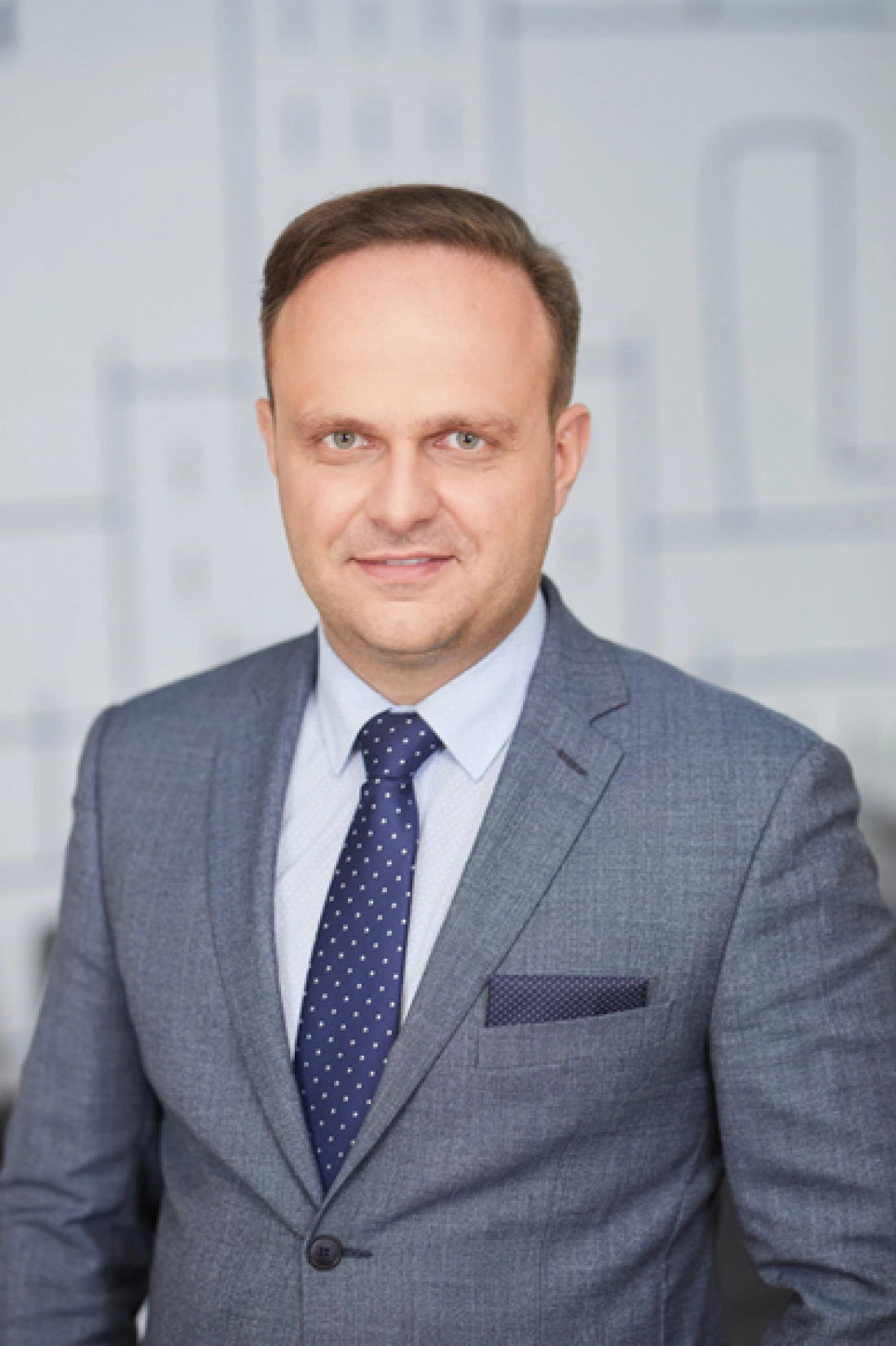 Mężczyzna w eleganckim garniturze, z uśmiechem, stojący na tle jasnej ściany.