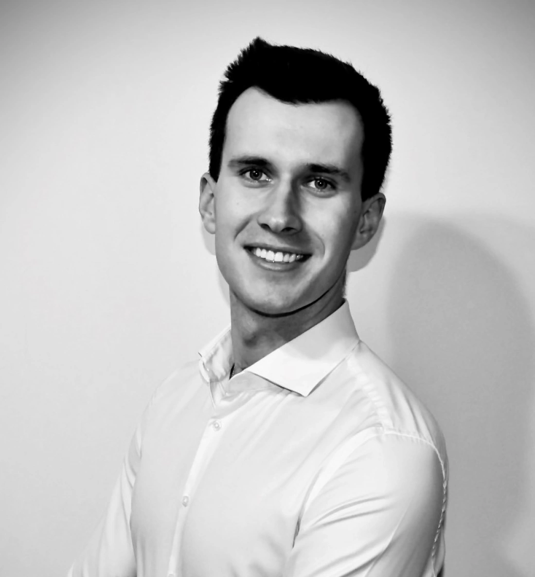 Mężczyzna w białej koszuli, uśmiechający się przy neutralnym tle.