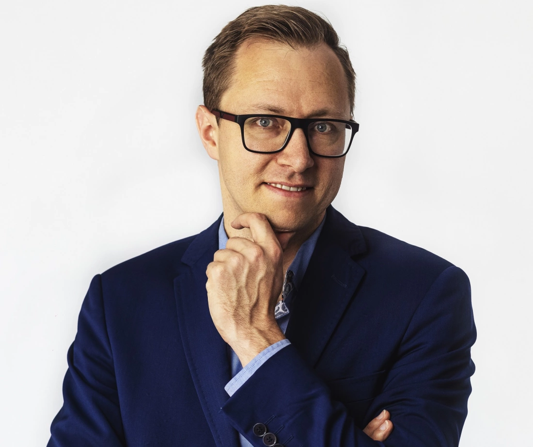 Mężczyzna w niebieskim garniturze z okularami, uśmiechający się i z ręką przy brodzie. Tło białe.