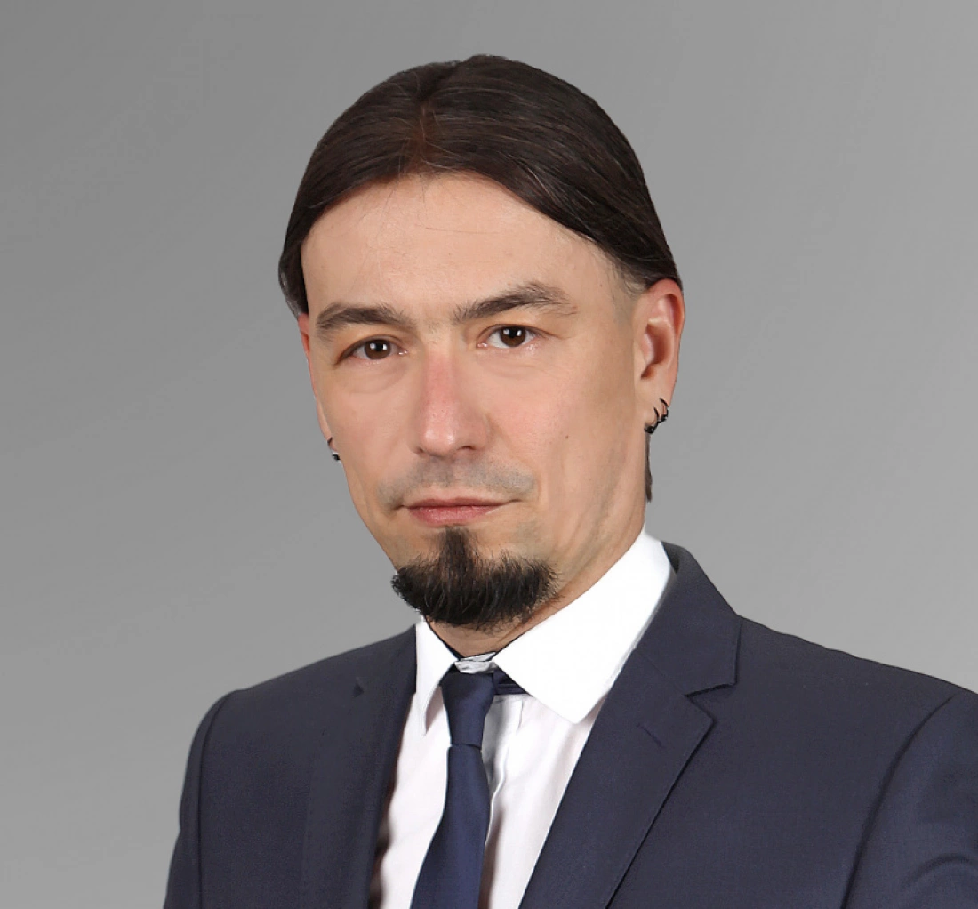 Mężczyzna w eleganckim garniturze z długimi włosami i kolczykiem, na neutralnym tle.