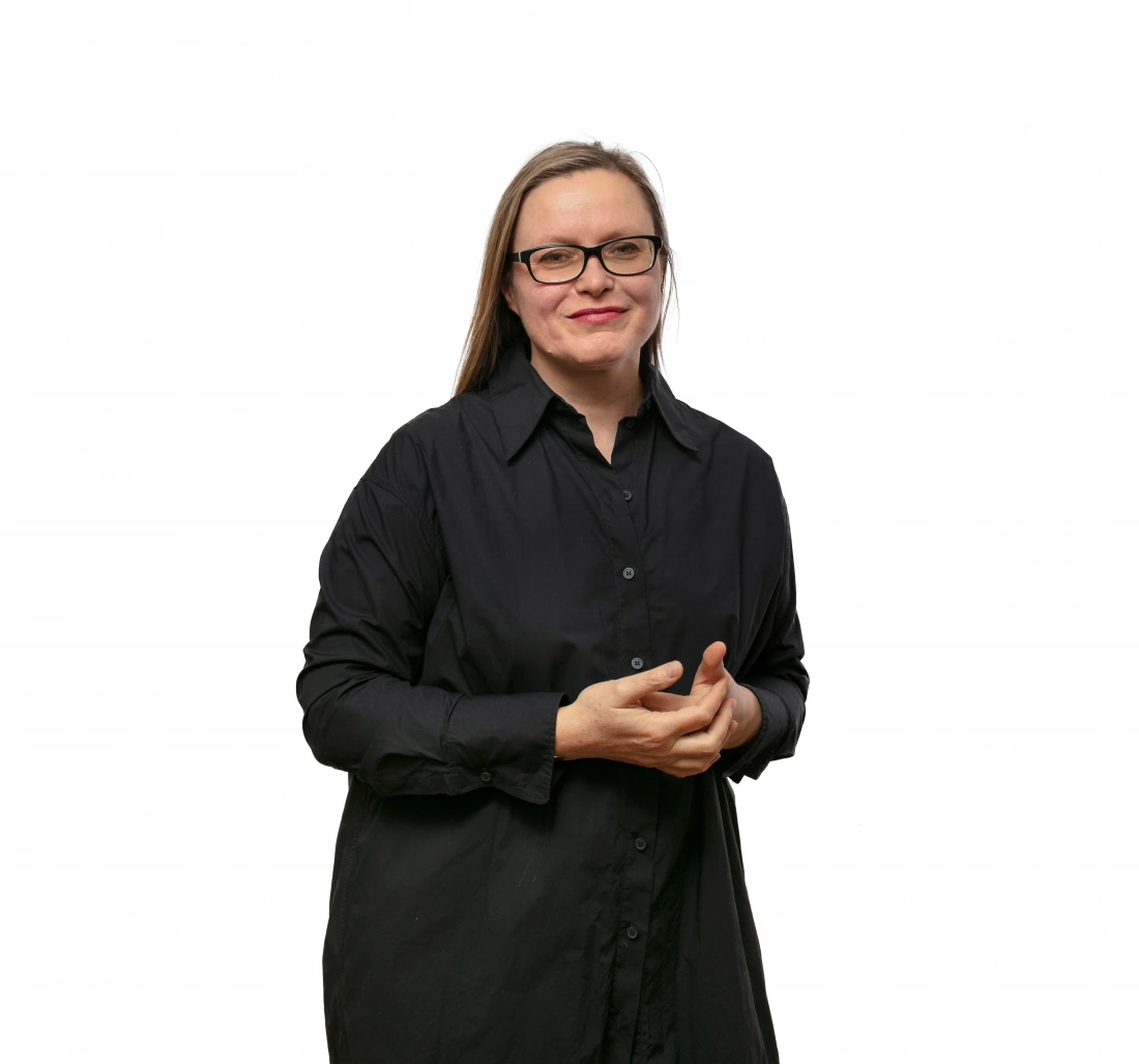 Kobieta w czarnej koszuli, z długimi włosami, uśmiechająca się i stojąca z rękami złożonymi przed sobą.