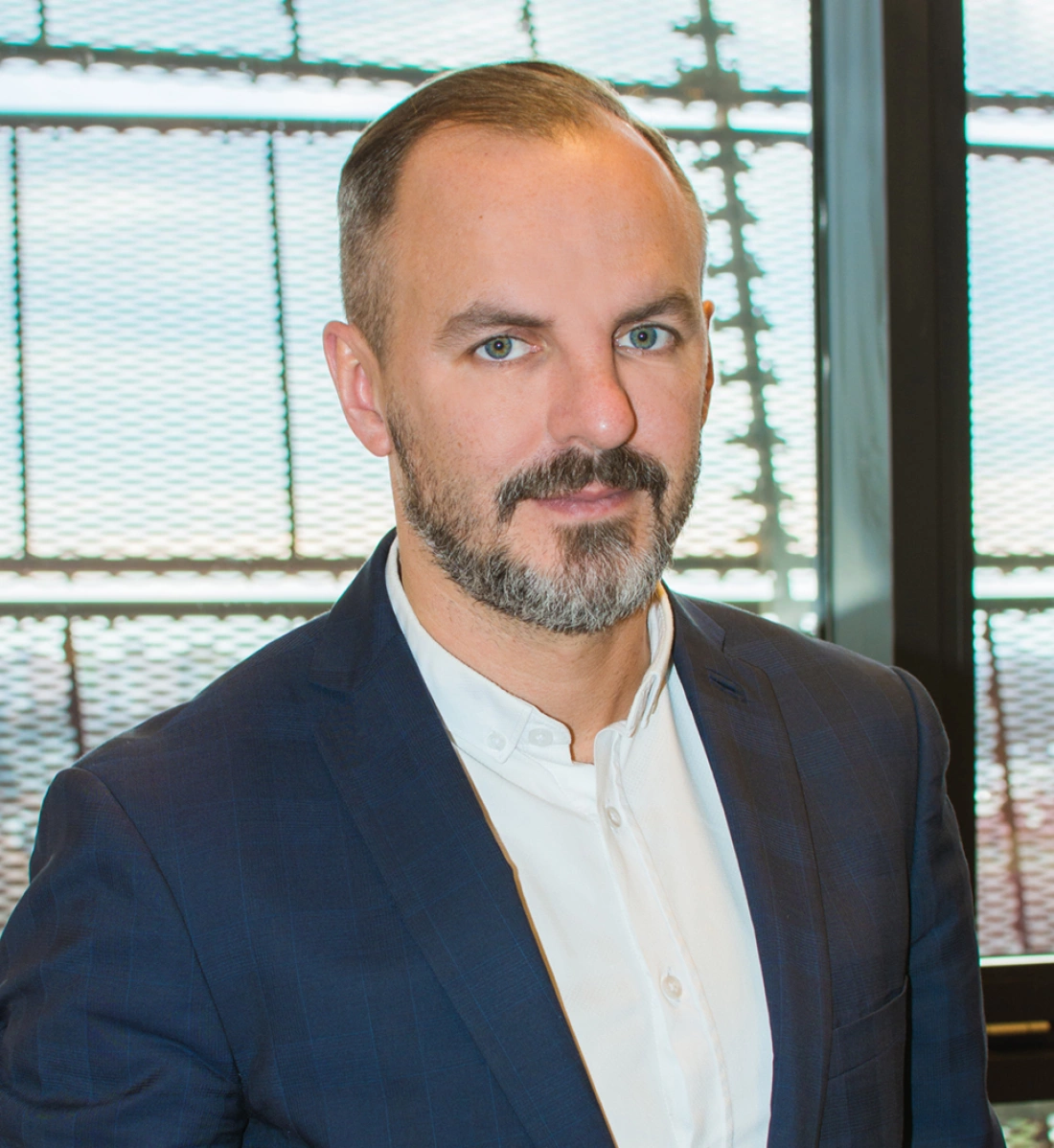 Mężczyzna w garniturze, z brodą, stoi w eleganckim otoczeniu.