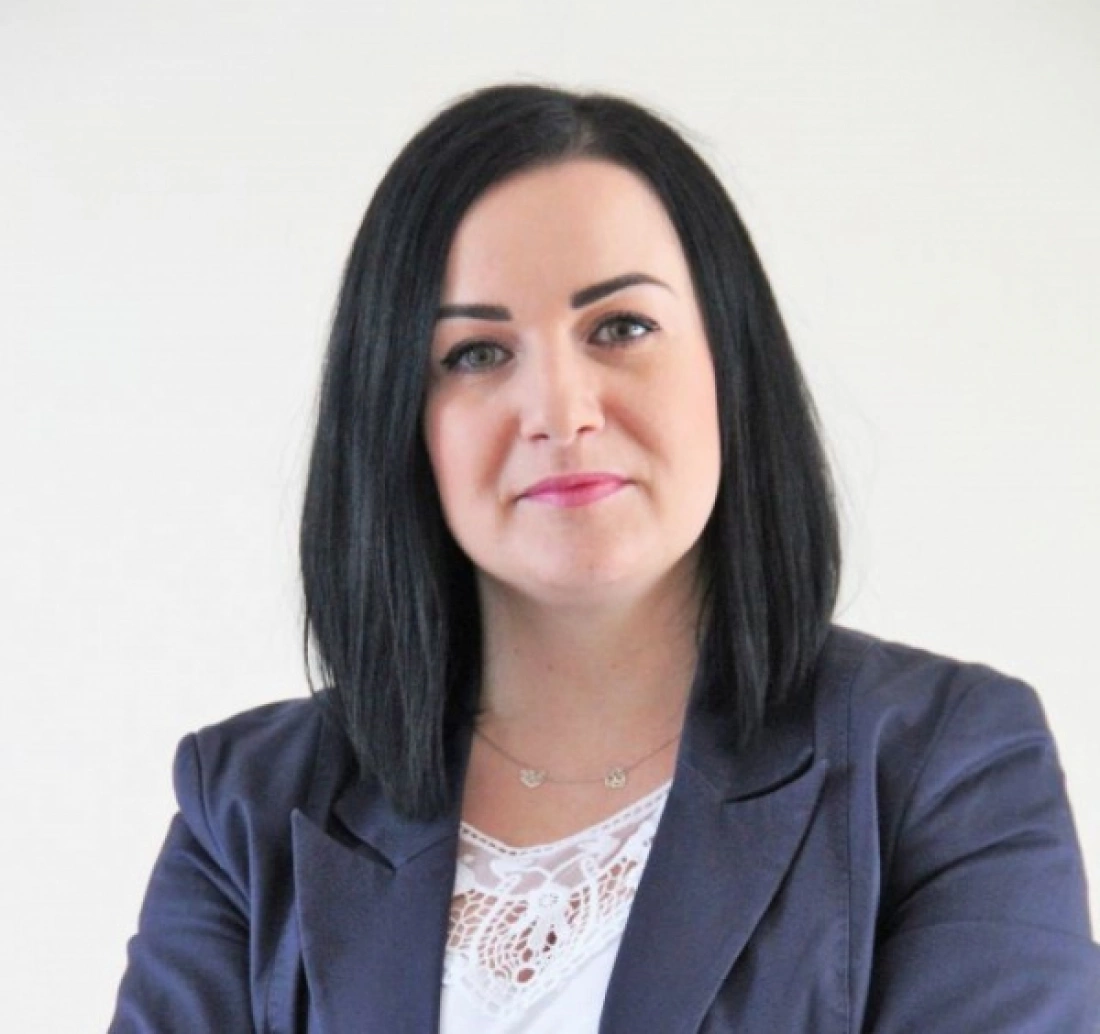 Kobieta z długimi, ciemnymi włosami, ubrana w granatową marynarkę, stoi z poważnym wyrazem twarzy.