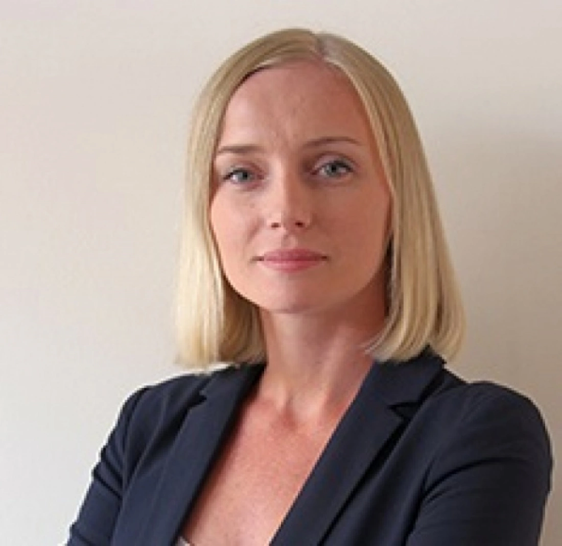 Kobieta o krótkich blond włosach w ciemnej marynarce, z poważnym wyrazem twarzy, stojąca na tle jasnej ściany.