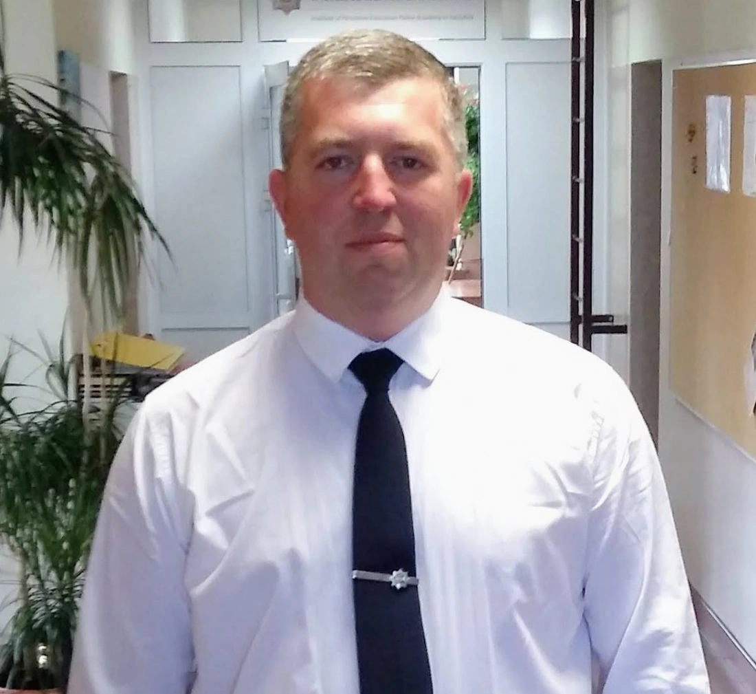 Mężczyzna w białej koszuli i czarnej krawacie stoi w korytarzu z roślinnymi dekoracjami.