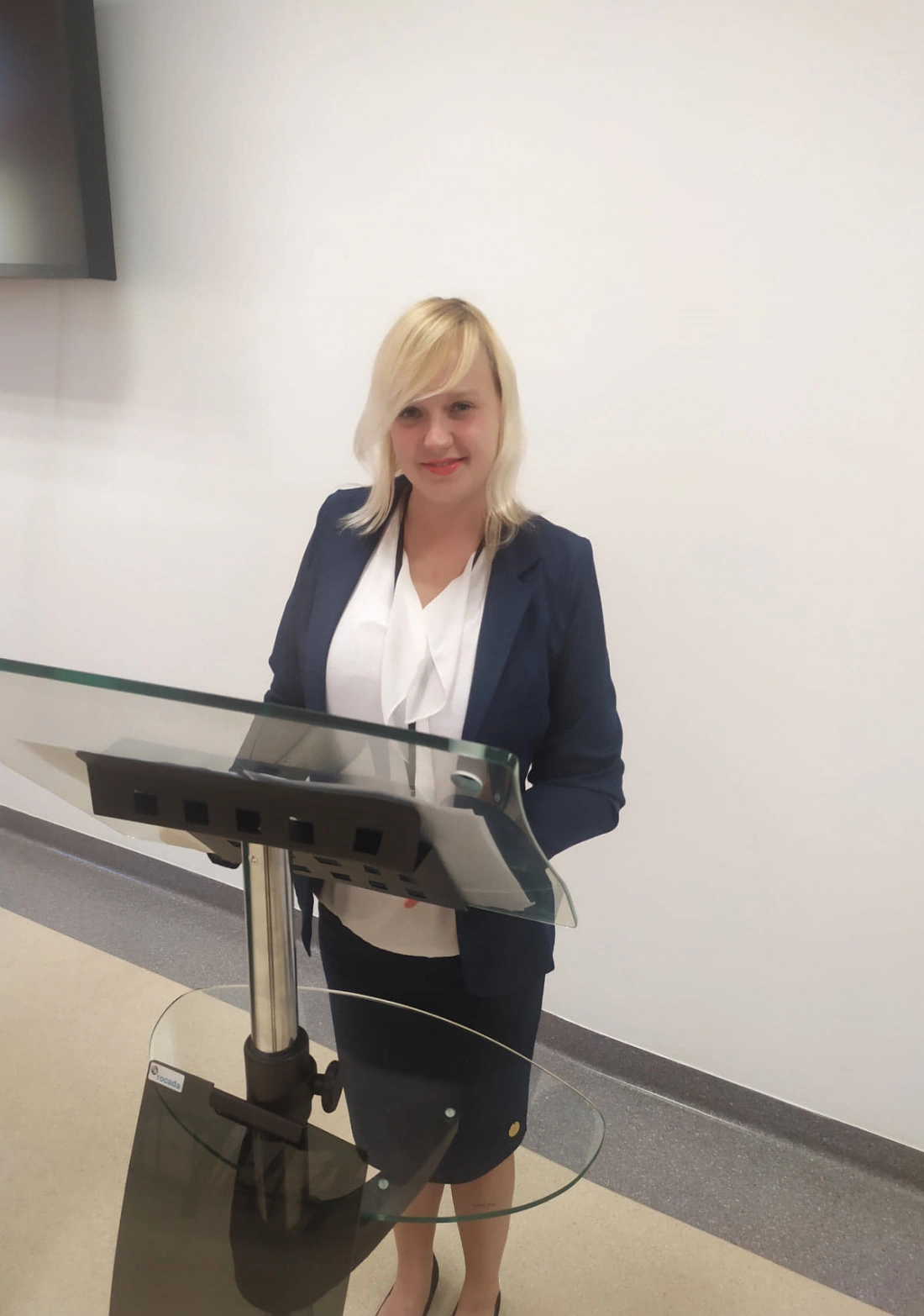 Kobieta w eleganckim stroju stojąca przy mównicy w jasnym pomieszczeniu.