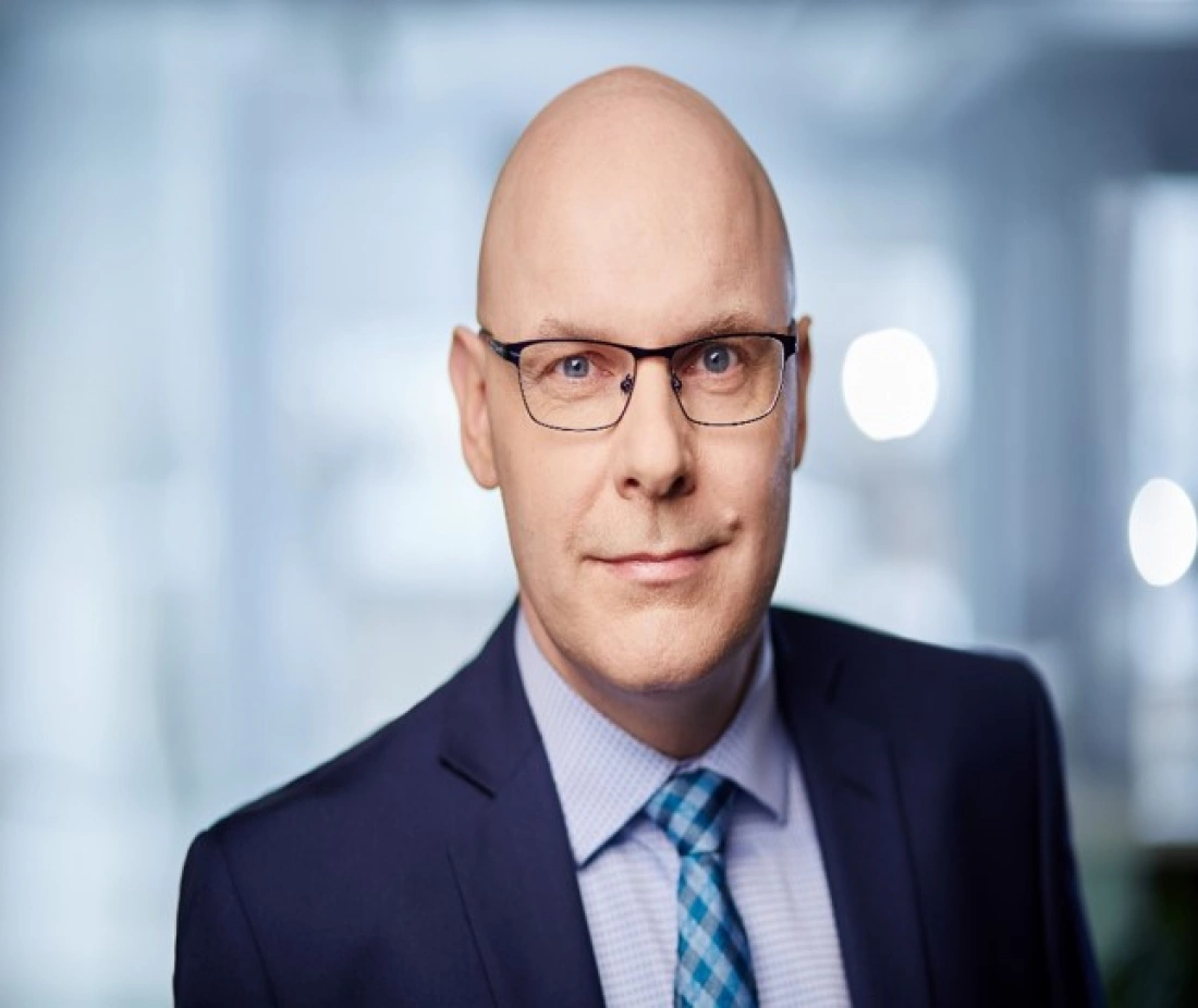 Mężczyzna w okularach, w garniturze, z lekko uśmiechniętą twarzą, na tle jasnego, rozmytego tła.