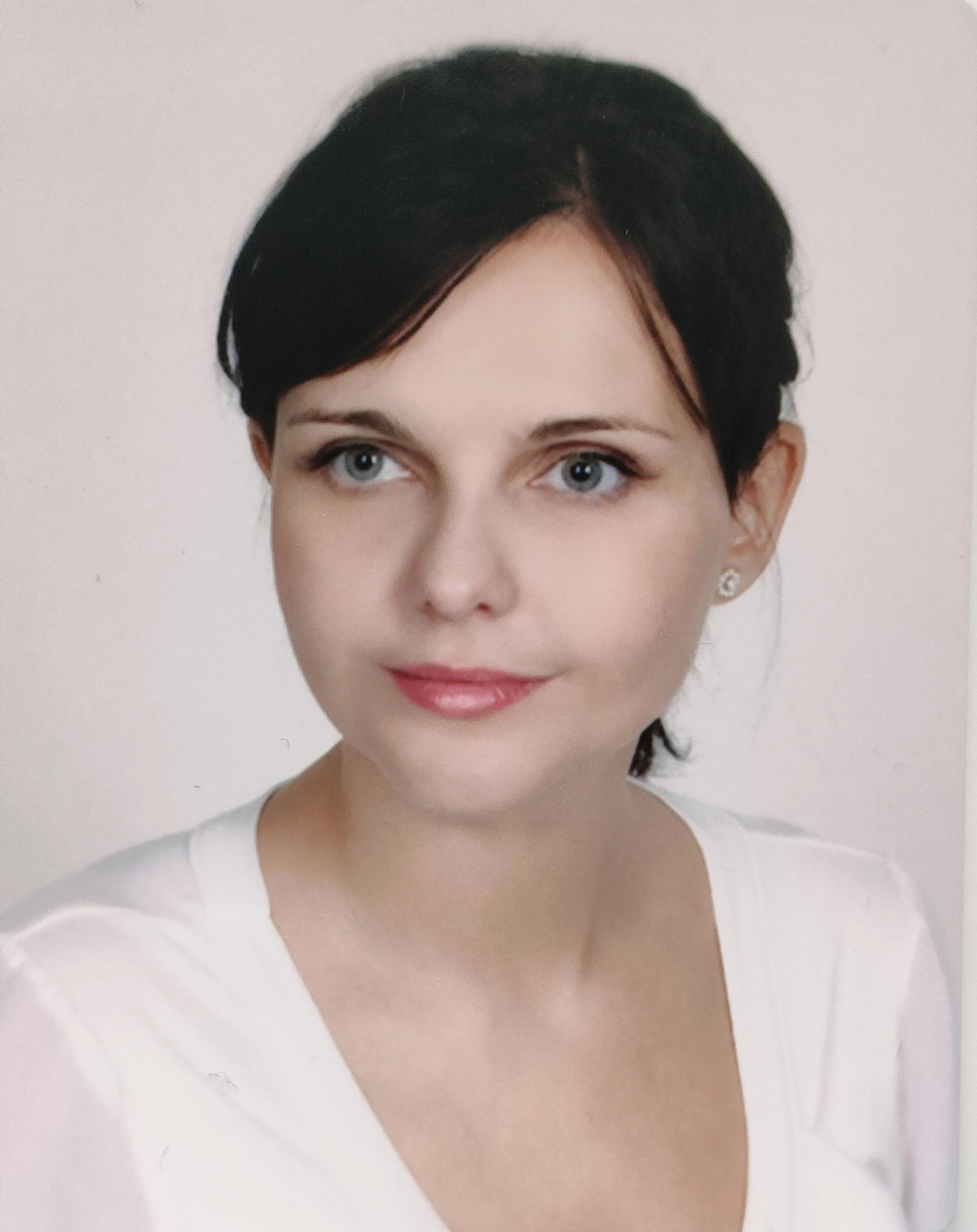 Portret młodej kobiety z błękitnymi oczami i ciemnymi włosami, uśmiechającej się subtelnie. Ubrana w białą bluzkę.