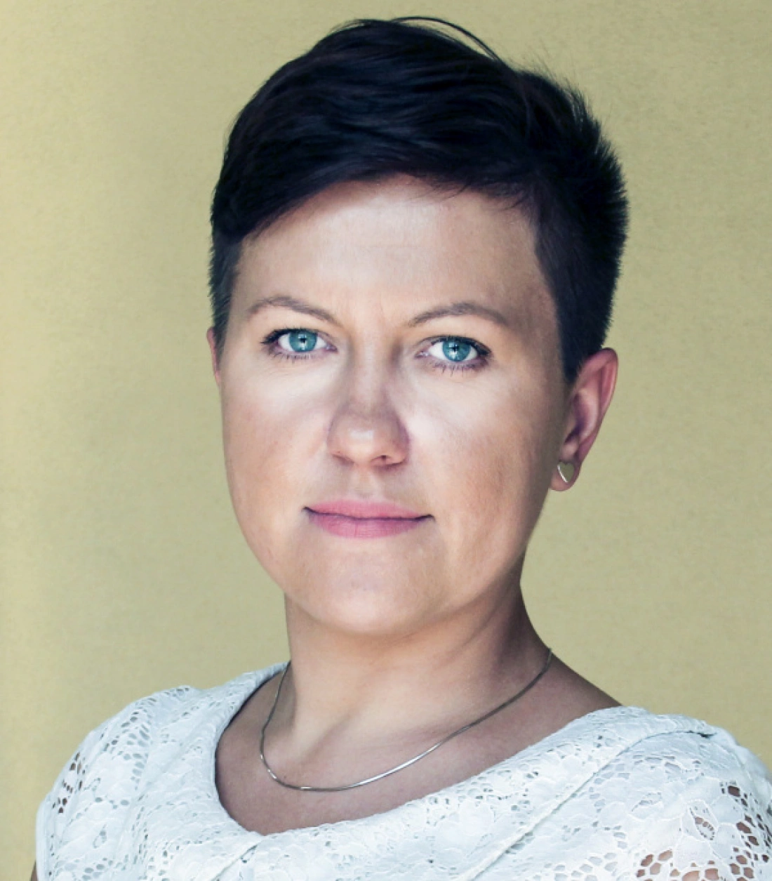 Portret kobiety o krótkich włosach, patrzącej prosto w obiektyw, z jasnym, neutralnym tłem.