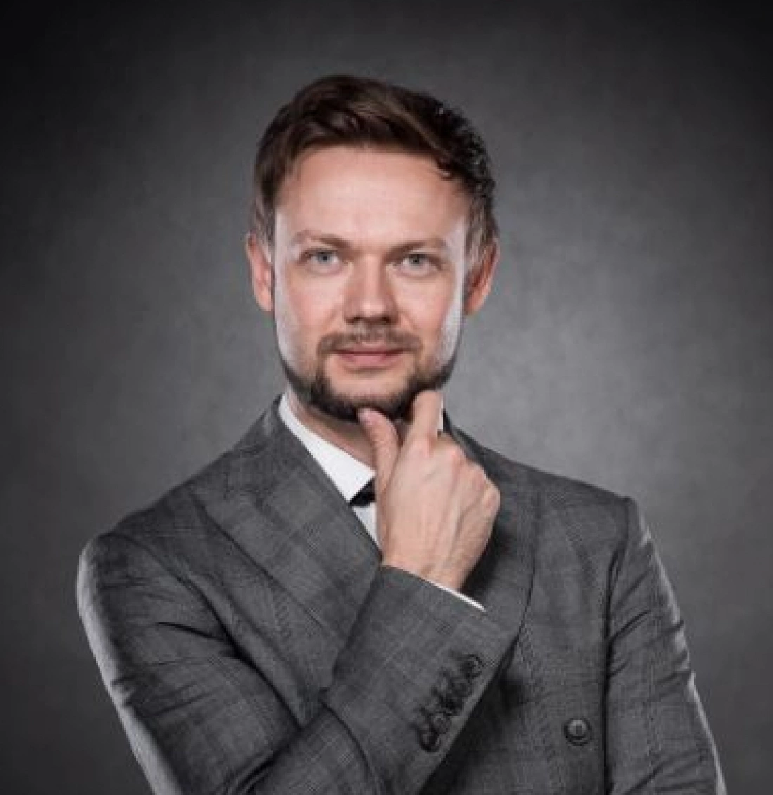 Mężczyzna w eleganckim garniturze, uśmiechający się i trzymający dłoń przy brodzie.