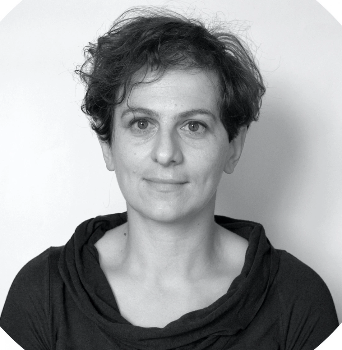 Portret osoby z krótkimi, kręconymi włosami, uśmiechającym się wyrazem twarzy, w czarnej bluzie. Tło jasne.