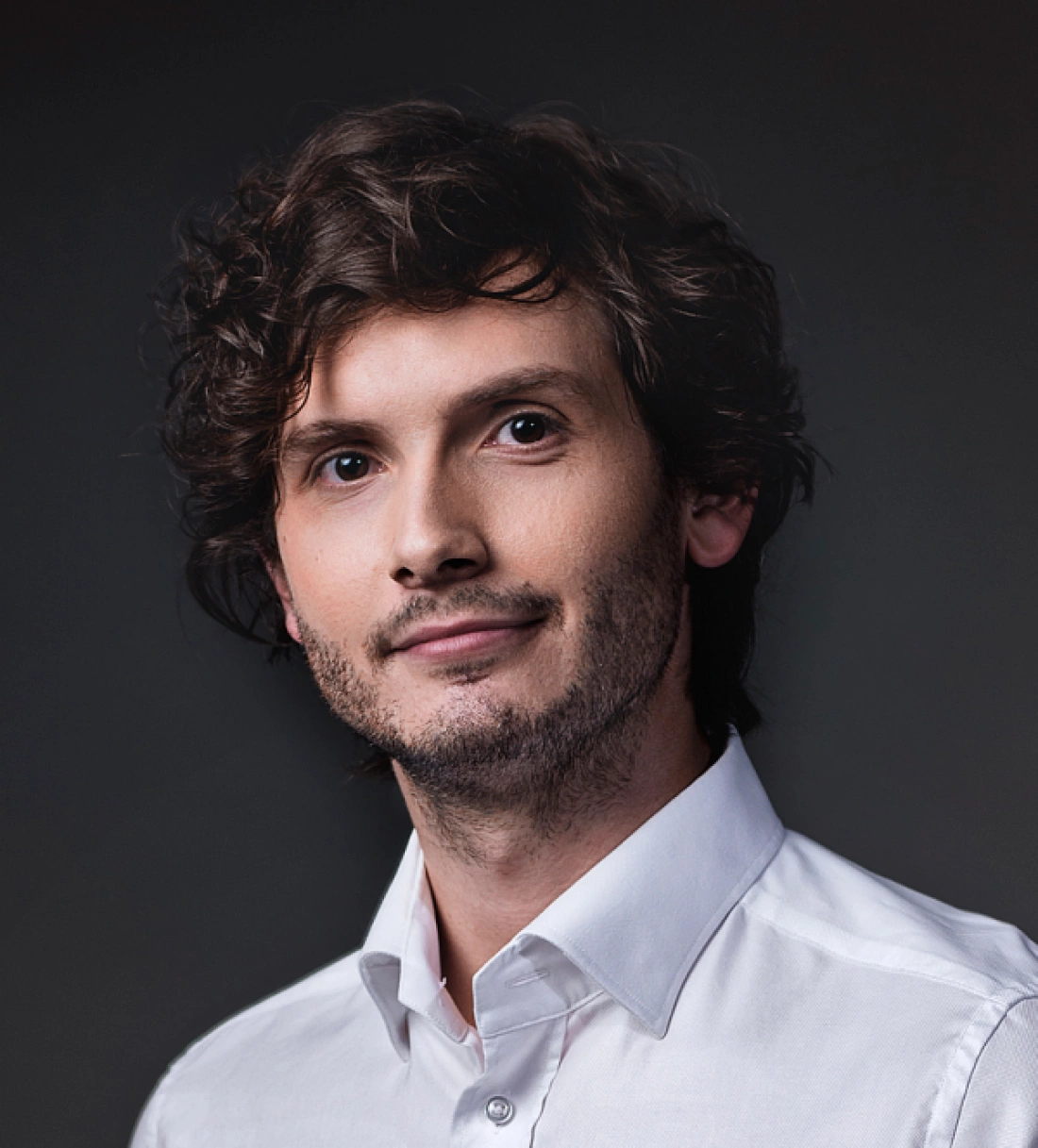 Portret mężczyzny z kręconymi włosami w białej koszuli, uśmiechającego się.