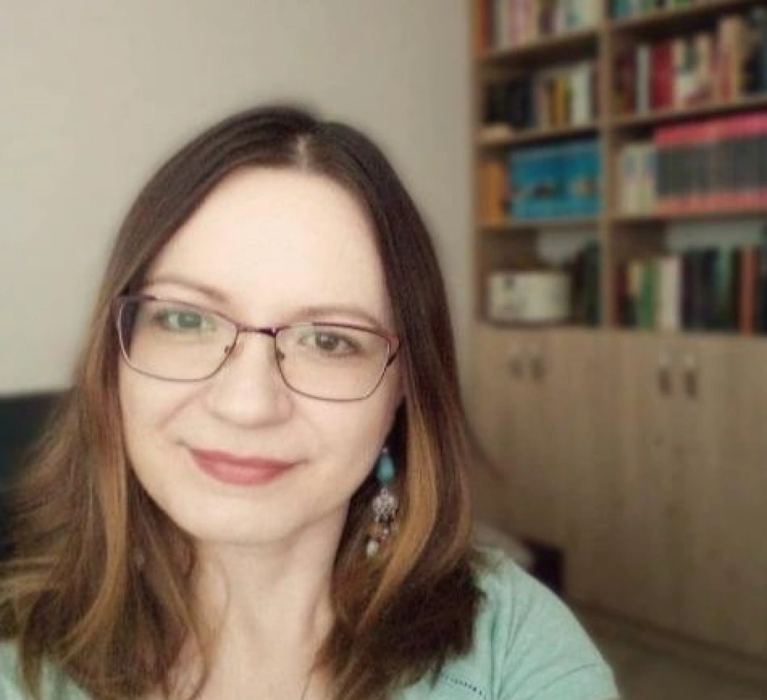 Kobieta z długimi włosami i okularami, uśmiechająca się przed regałem z książkami.