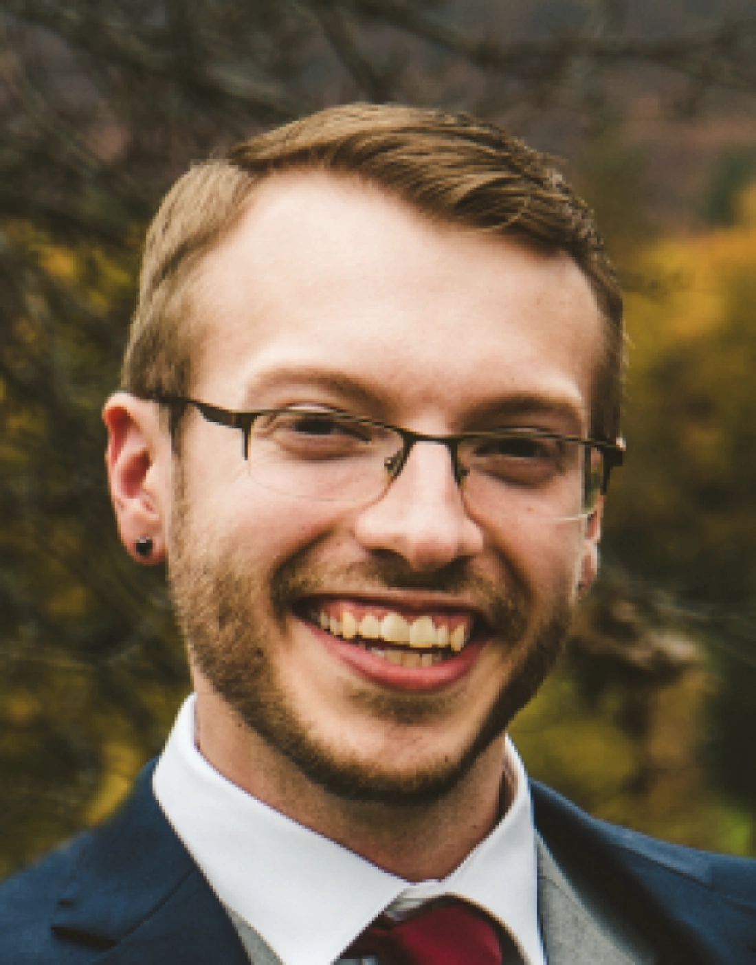 Mężczyzna w garniturze uśmiecha się, stojąc na tle jesiennego krajobrazu.