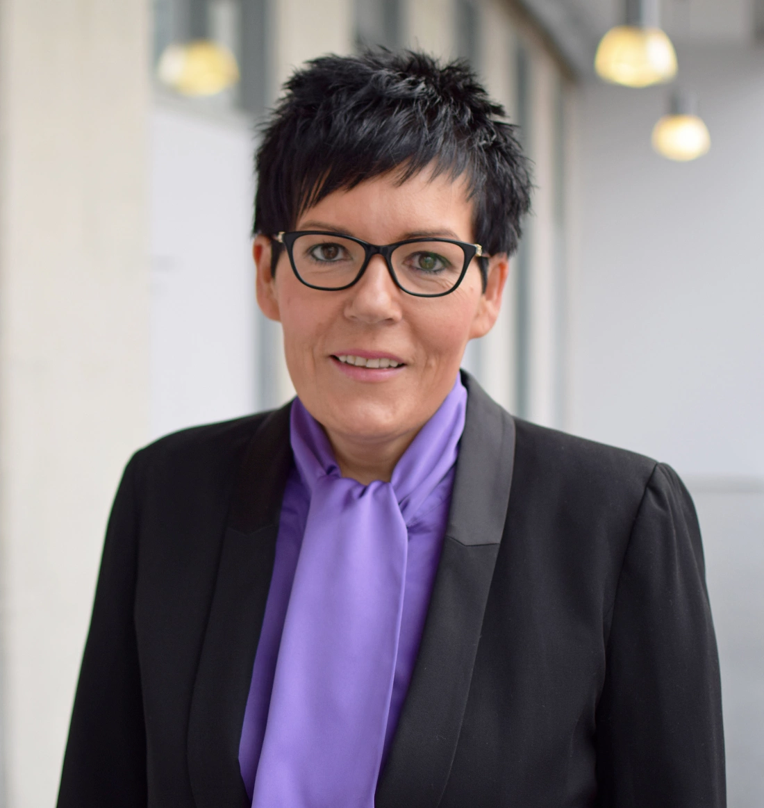 Kobieta w czarnym garniturze i fioletowej apaszce, uśmiechająca się.