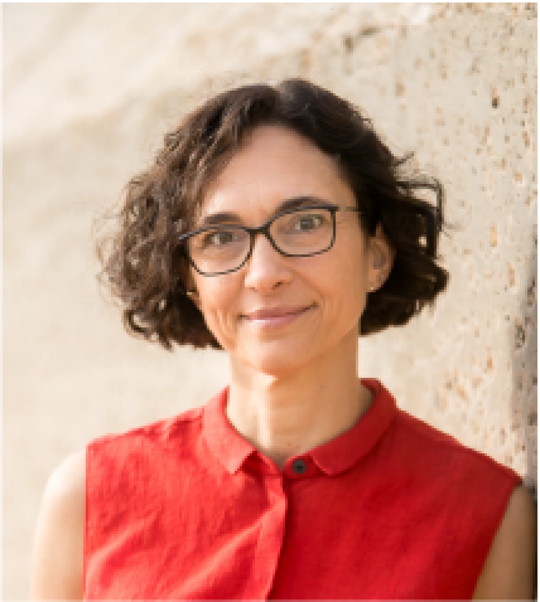 Kobieta w okularach, uśmiechająca się na tle ściany. Ma krótkie, kręcone włosy i na sobie czerwoną koszulę.