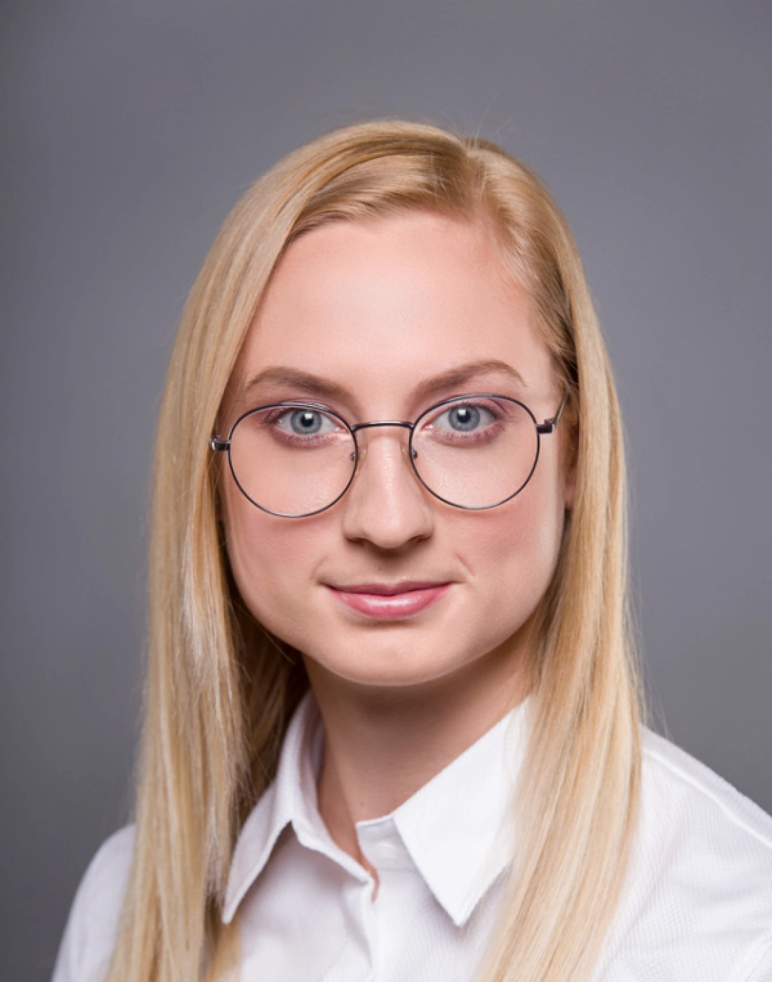 Portret osoby z długimi blond włosami, noszącej okrągłe okulary, w białej koszuli na tle szarym.