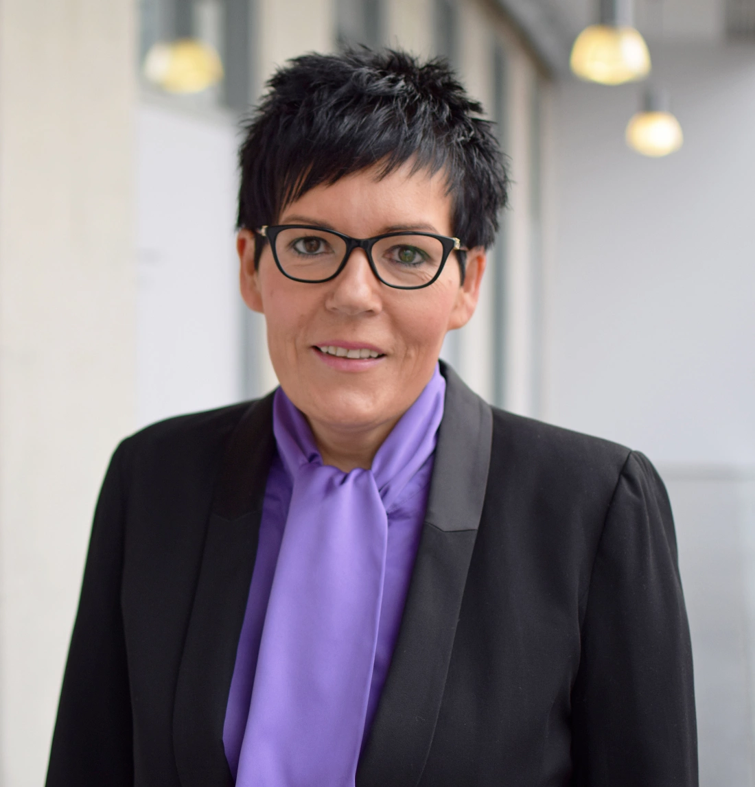 Kobieta w eleganckim stroju z krótkimi ciemnymi włosami i okularami, uśmiechająca się.