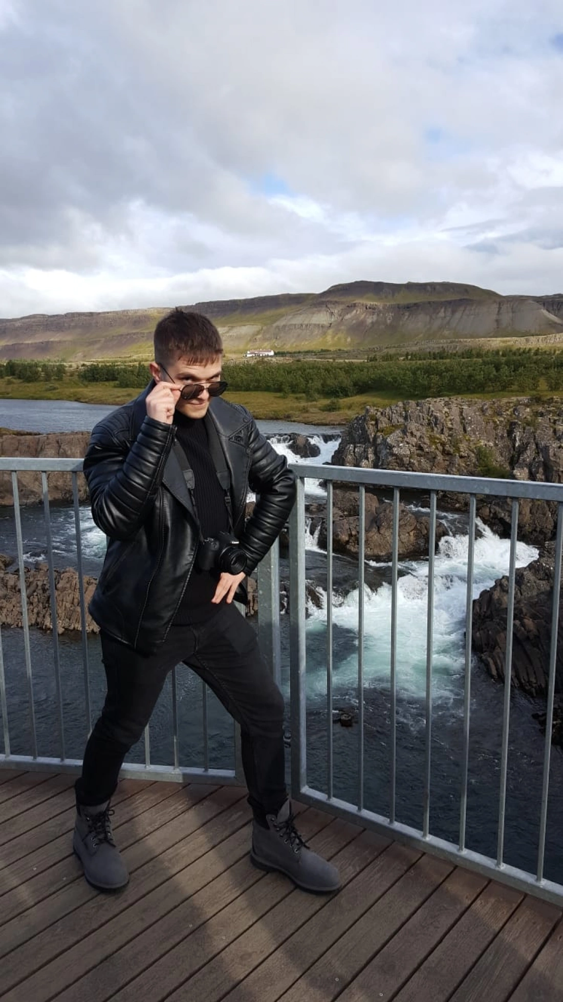 Mężczyzna w skórzanej kurtce stoi na drewnianym tarasie z widokiem na wodospad i góry.