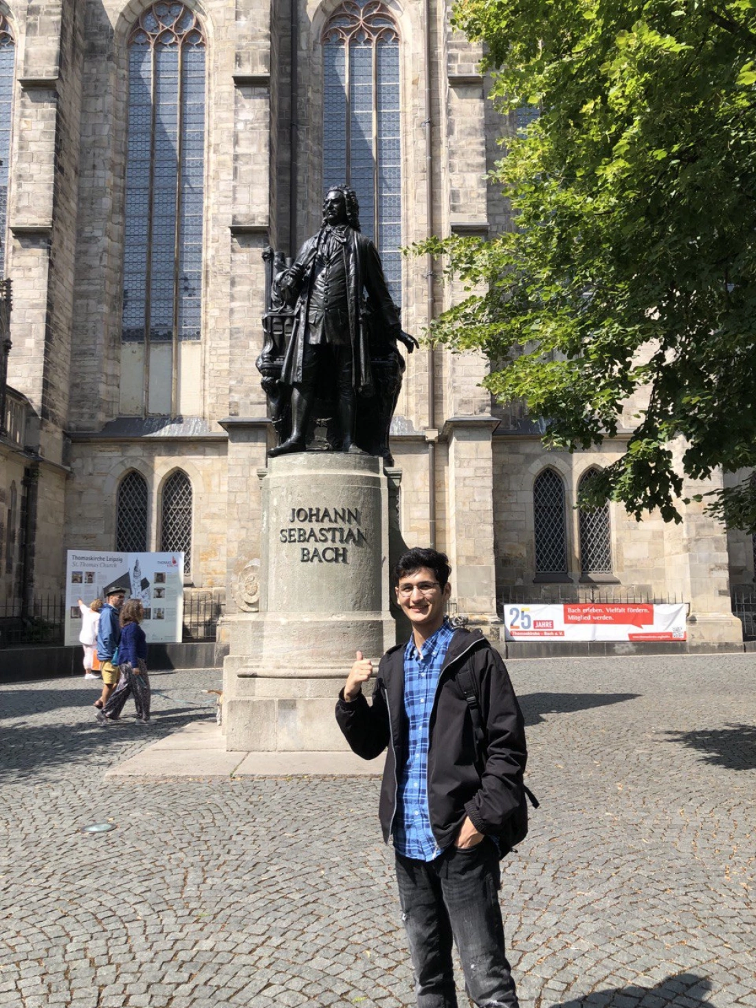 Młody mężczyzna stoi obok pomnika Johanna Sebastiana Bacha, z uśmiechem i kciukiem w górze, w tle zabytkowy budynek.