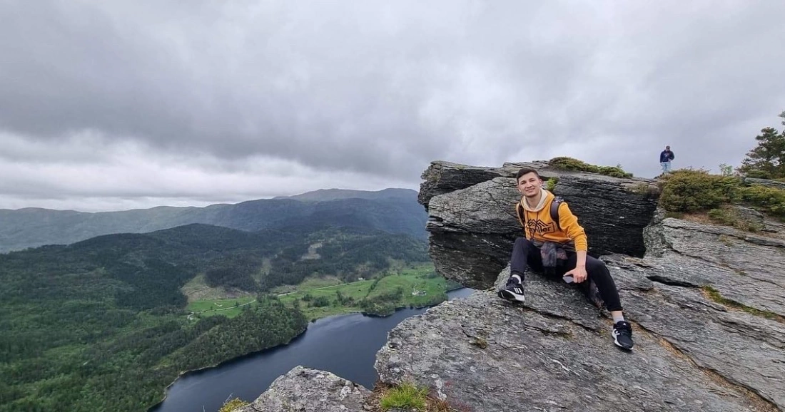 Mężczyzna siedzi na wystającym głazie z widokiem na zieloną dolinę i jezioro w tle.