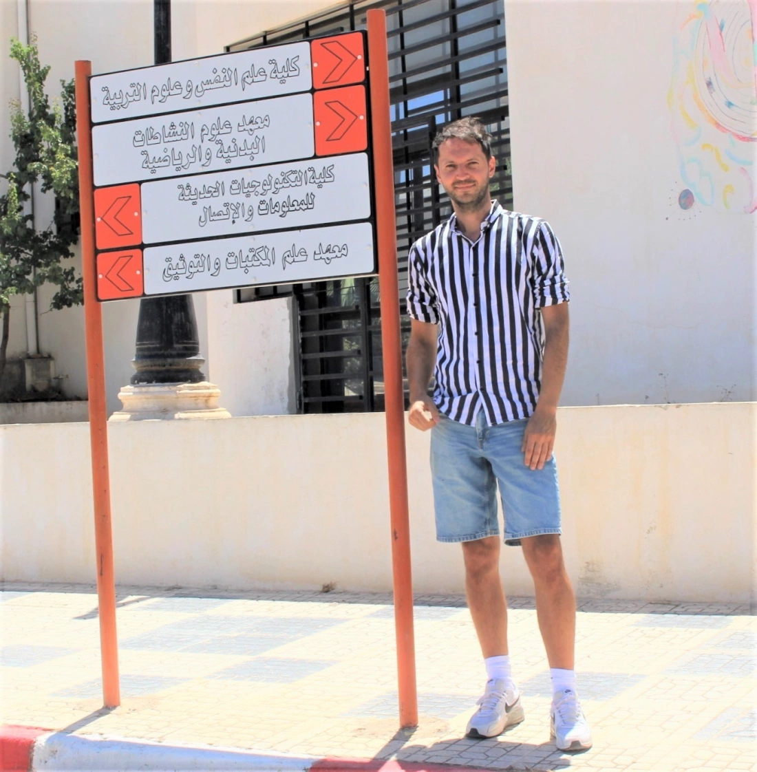 Mężczyzna w pasiastym kolorze t-shirtu stoi obok tablicy informacyjnej na tle budynku.