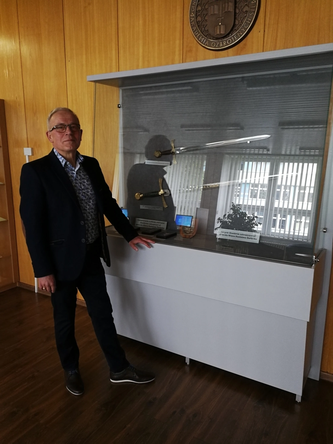 Mężczyzna stoi obok witryny z eksponatami, w tym mieczami, w pomieszczeniu z drewnianymi ścianami.