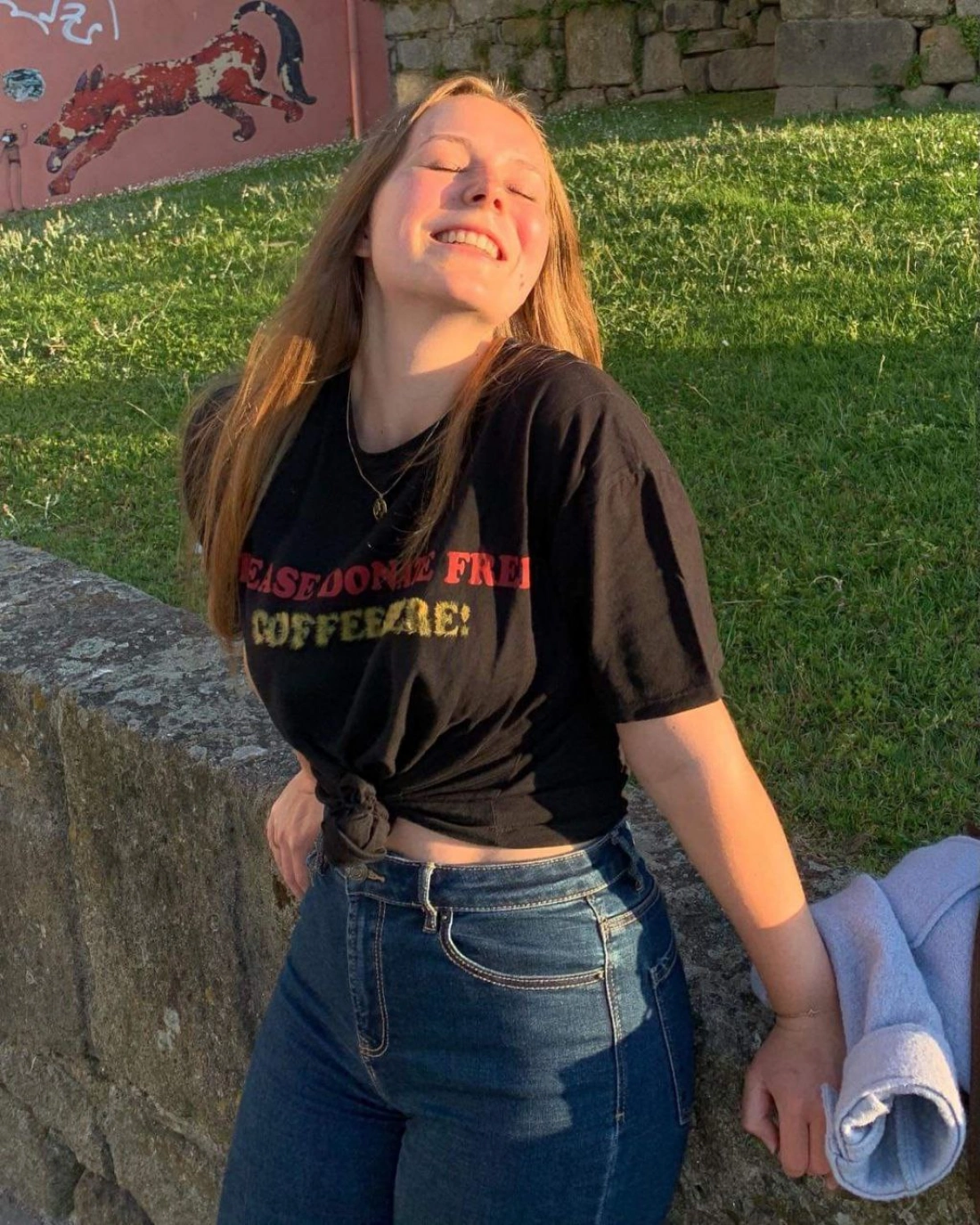Młoda osoba uśmiechająca się na tle trawy i kamieni, w czarnej koszulce z napisem.
