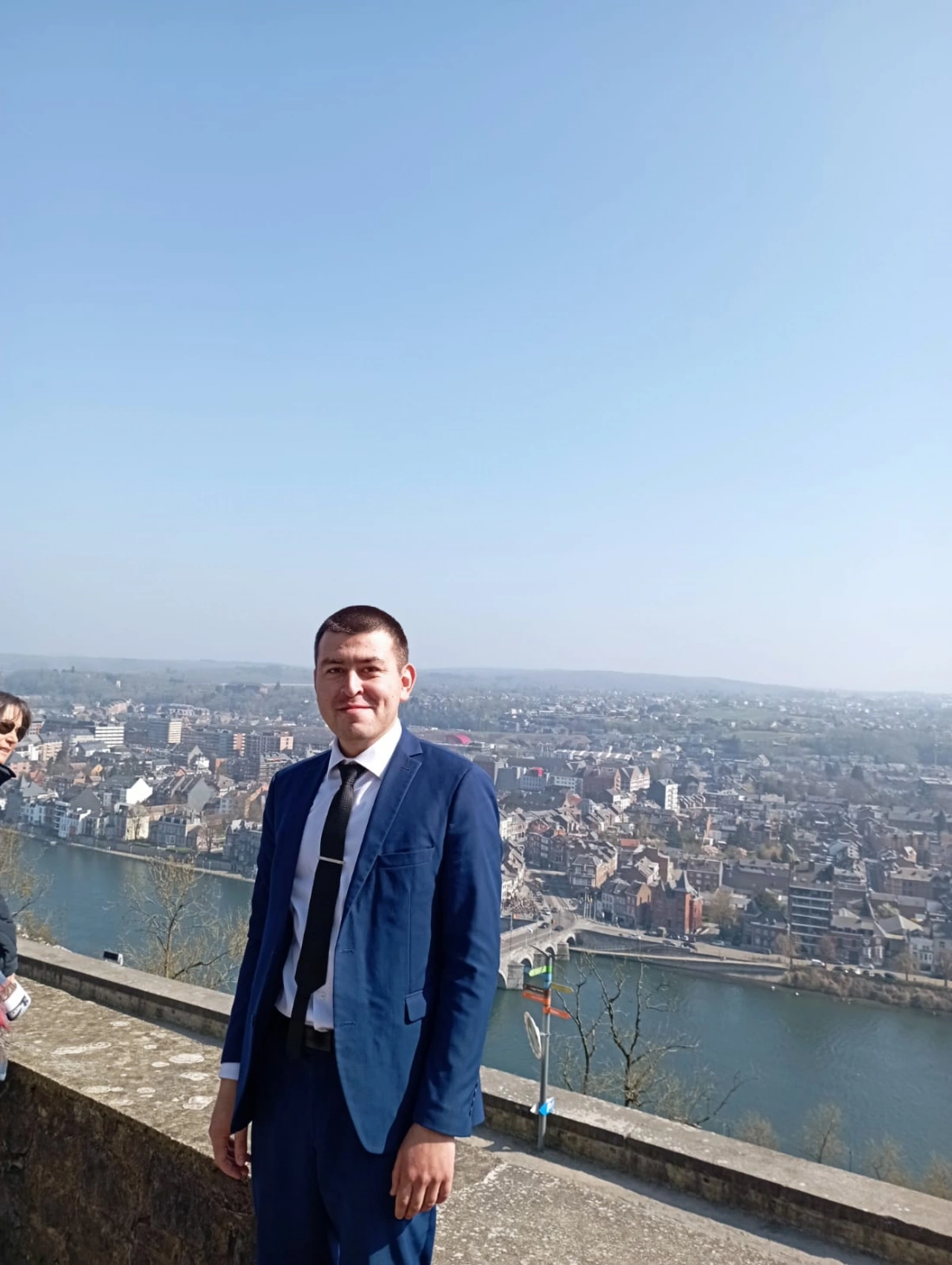 Mężczyzna w garniturze stoi na tle miasta z widokiem na rzekę i niebo.