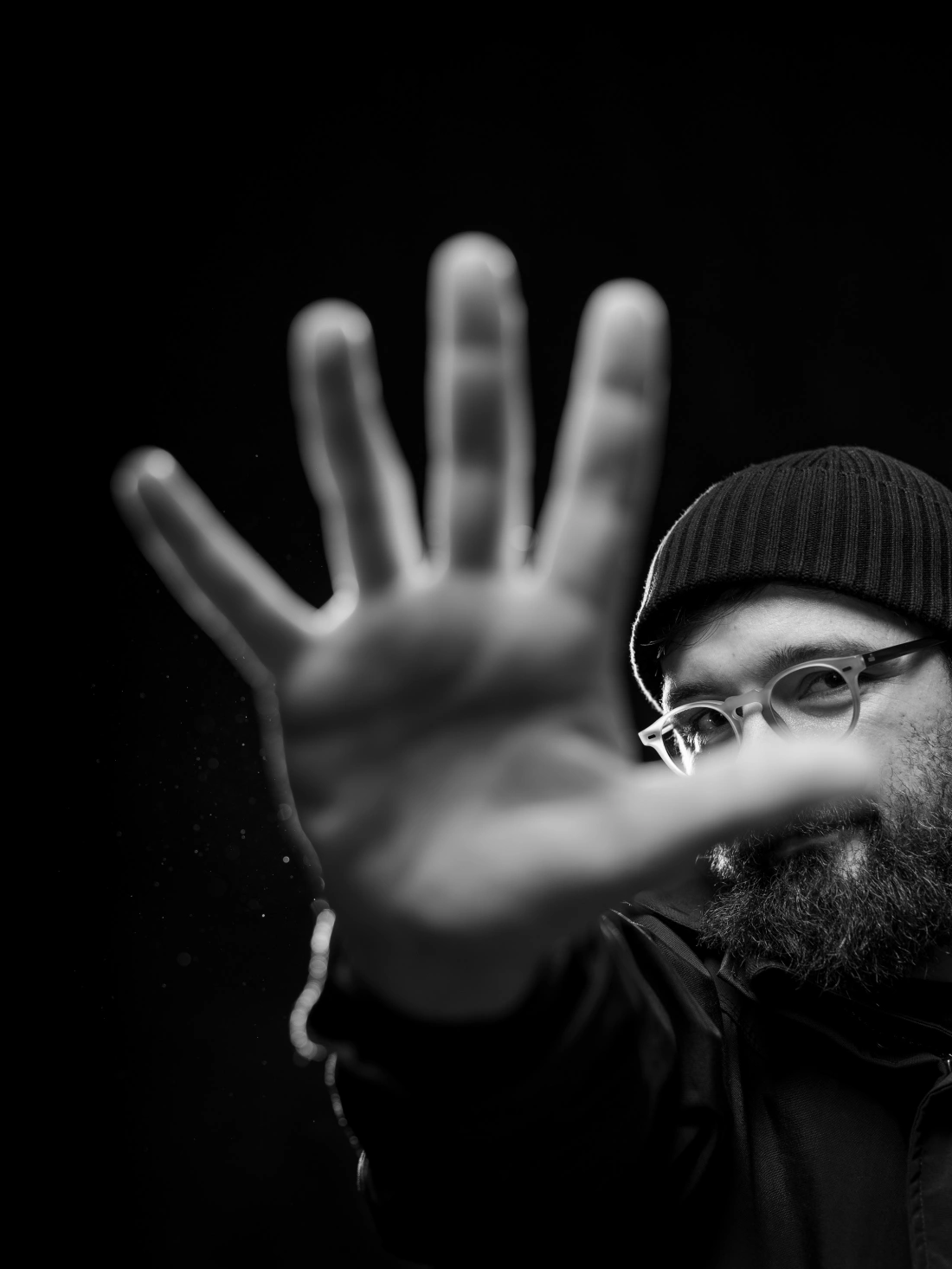 Mężczyzna w czapce z długą brodą, odsłoniętą dłonią kierującą w stronę kamery, na tle czarnym.
