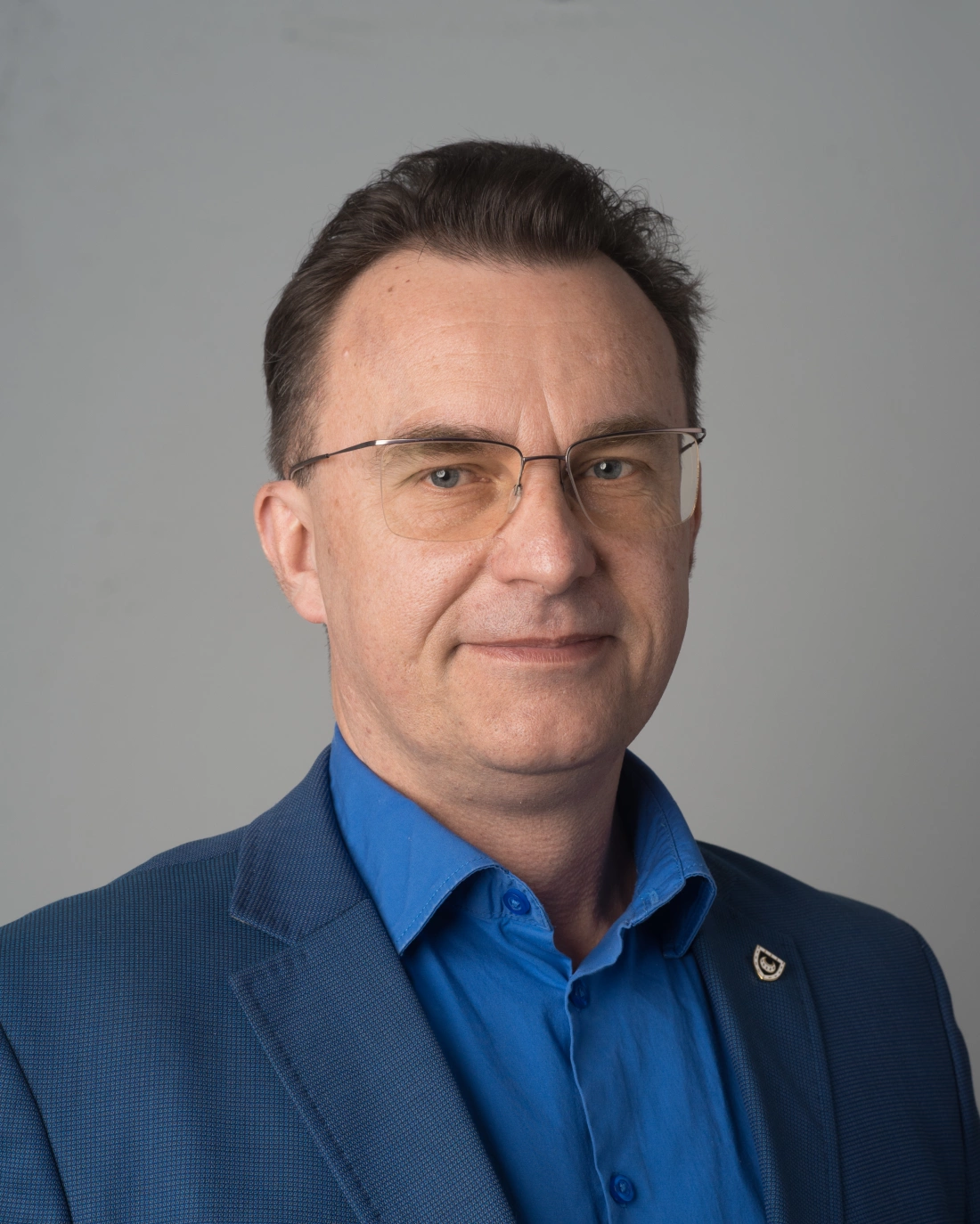 Ujęcie mężczyzny w garniturze, z okularami, na tle jednolitej ściany.