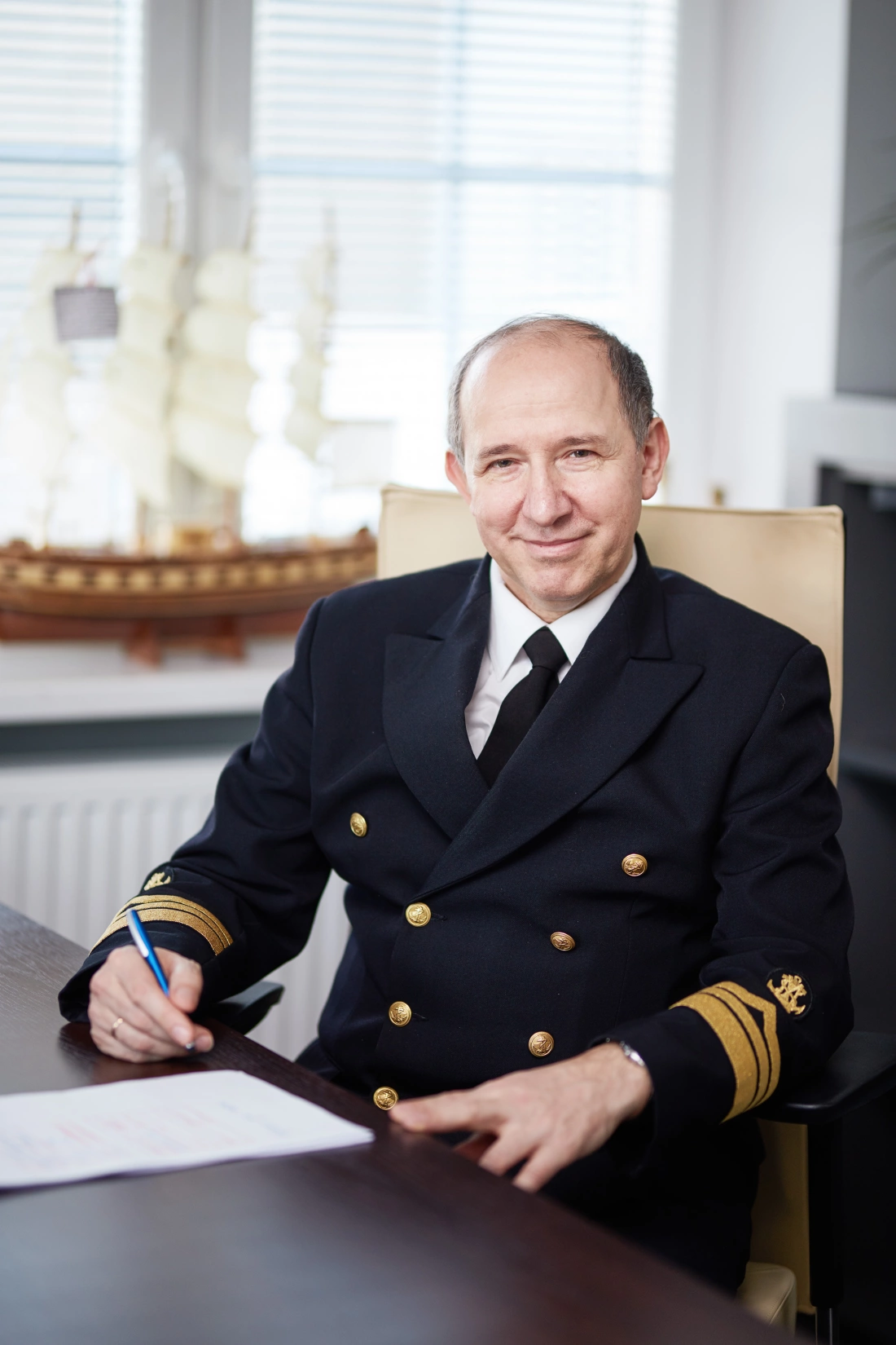 Mężczyzna w mundurze marynarskim siedzi przy biurku, pisząc notatki. W tle widoczny model statku.