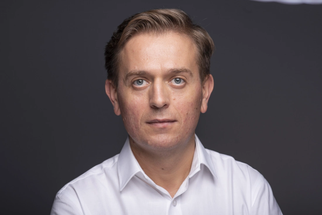 Mężczyzna w białej koszuli, patrzący w kamerę na ciemnym tle.