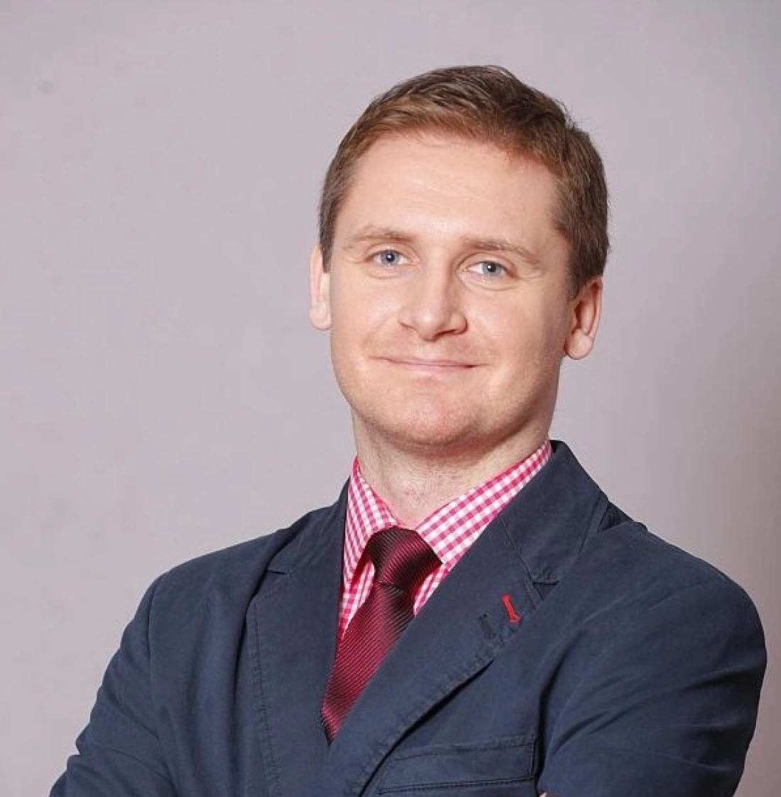 Mężczyzna w eleganckim ubraniu, z uśmiechem na twarzy, pozujący z założonymi rękami.