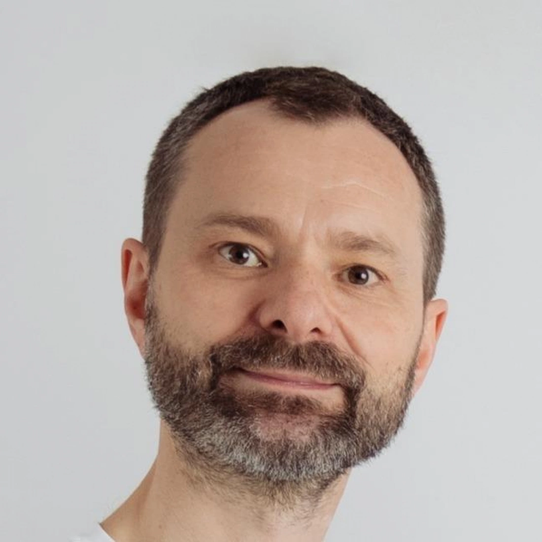 Mężczyzna z brodą uśmiecha się, stojąc na jasnym tle.