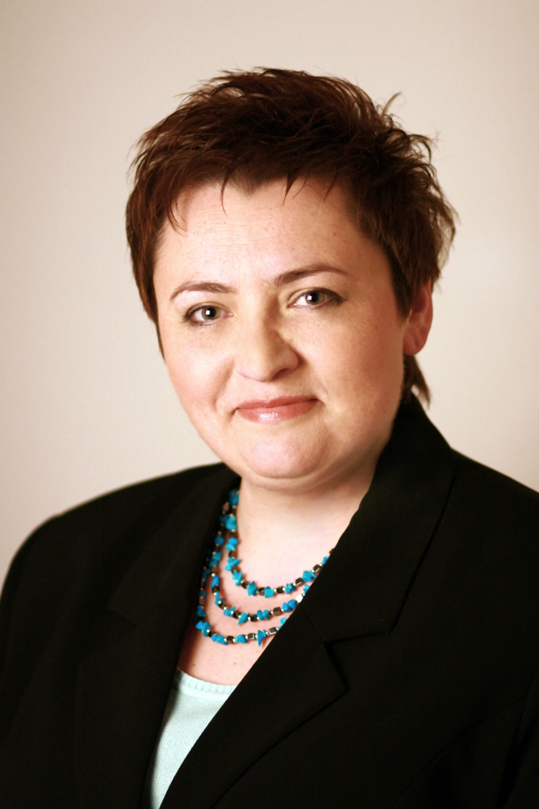 Kobieta w garniturze z krótkimi włosami i naszyjnikiem, uśmiechająca się na tle jasnego tła.