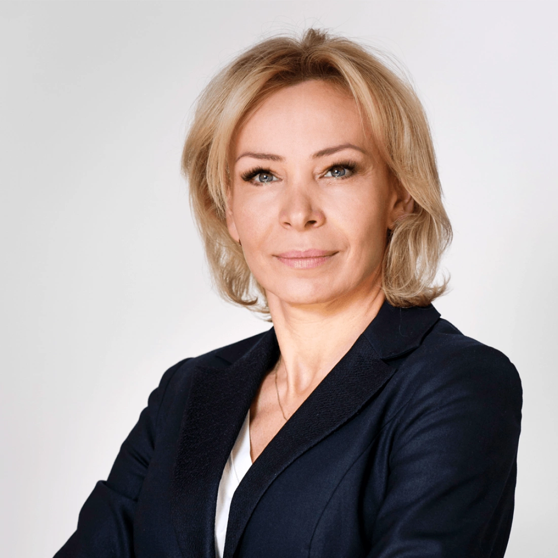 Kobieta w eleganckim stroju, z delikatnym uśmiechem, stoi z założonymi rękami na tle jasnej ściany.