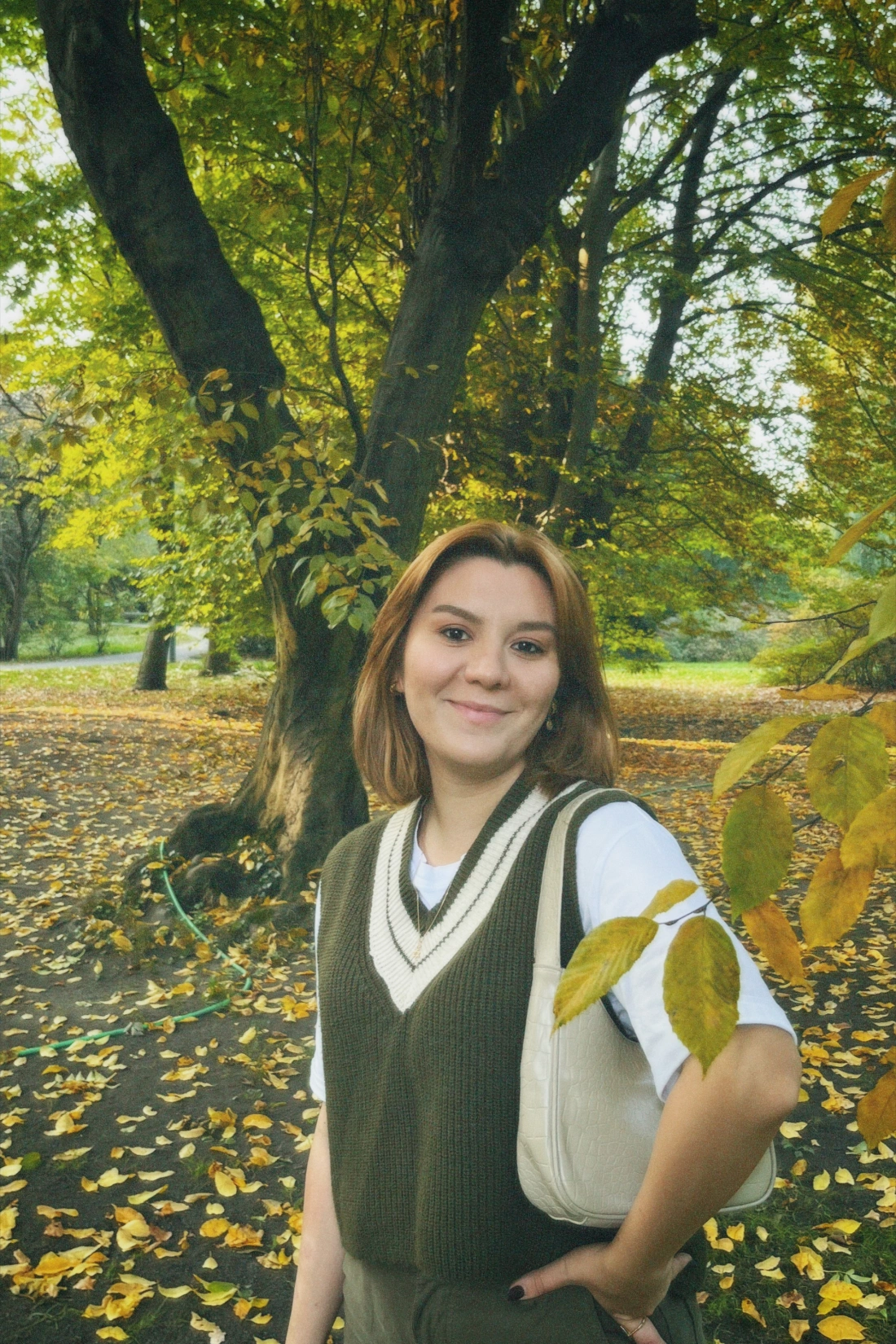 Młoda kobieta stoi w parku wśród żółknących liści drzew, uśmiechając się do aparatu.