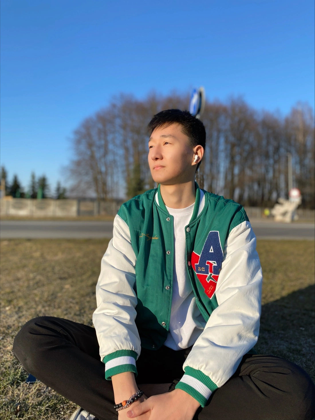 Młody mężczyzna w zimowej kurtce siedzi na trawie w słoneczny dzień, patrząc w stronę nieba.