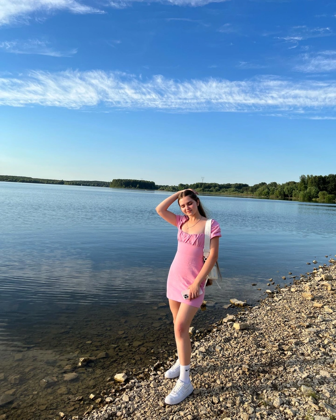 Młoda kobieta w różowej sukience stoi nad brzegiem jeziora, z błękitnym niebem w tle.