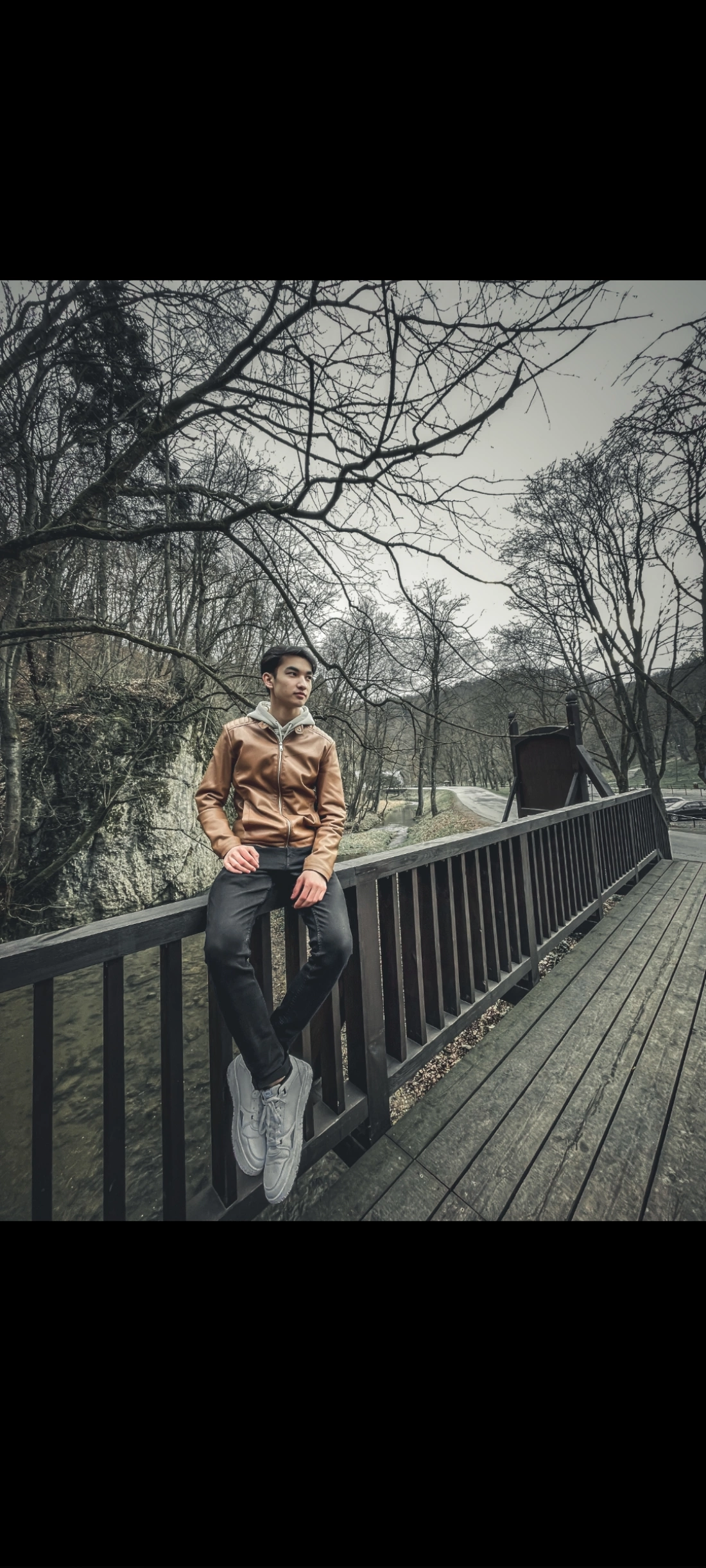 Młody mężczyzna w brązowej kurtce siedzi na moście w lesie.