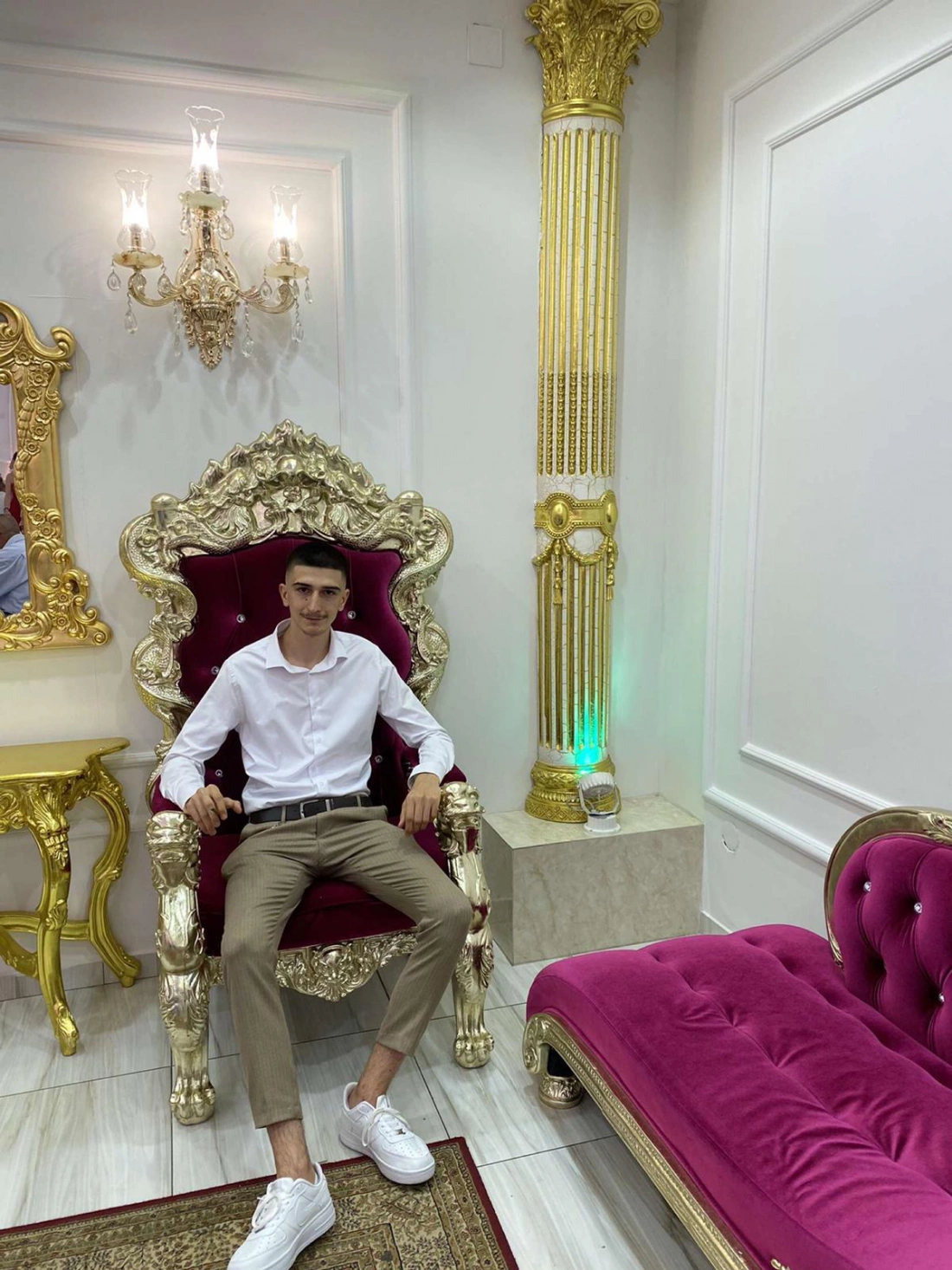 Młody mężczyzna w białej koszuli siedzi na ozdobnym, złotym fotelu w eleganckim wnętrzu. Obok niego znajduje się różowa kanapa.