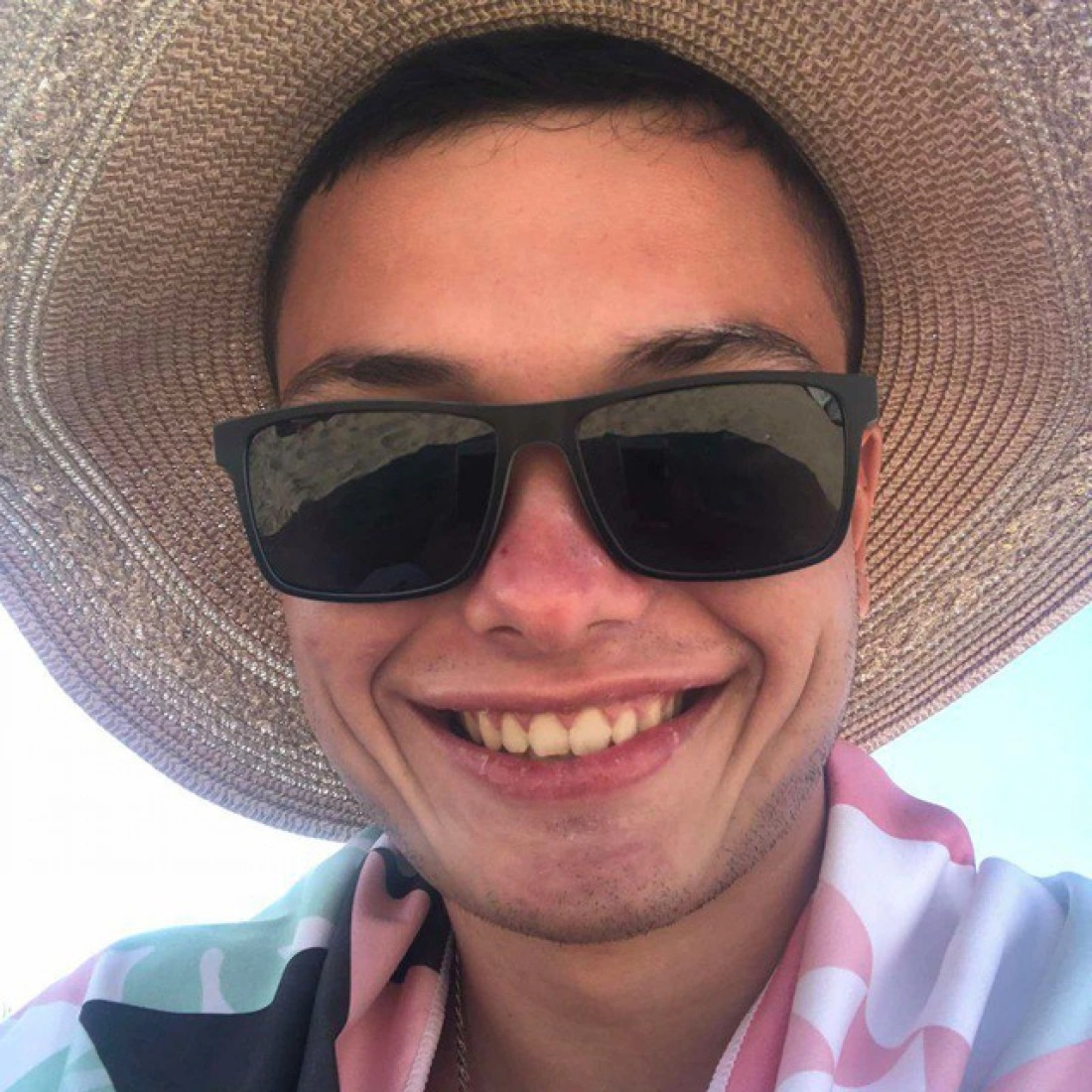 Mężczyzna w dużym kapeluszu i okularach przeciwsłonecznych uśmiecha się na tle plaży.