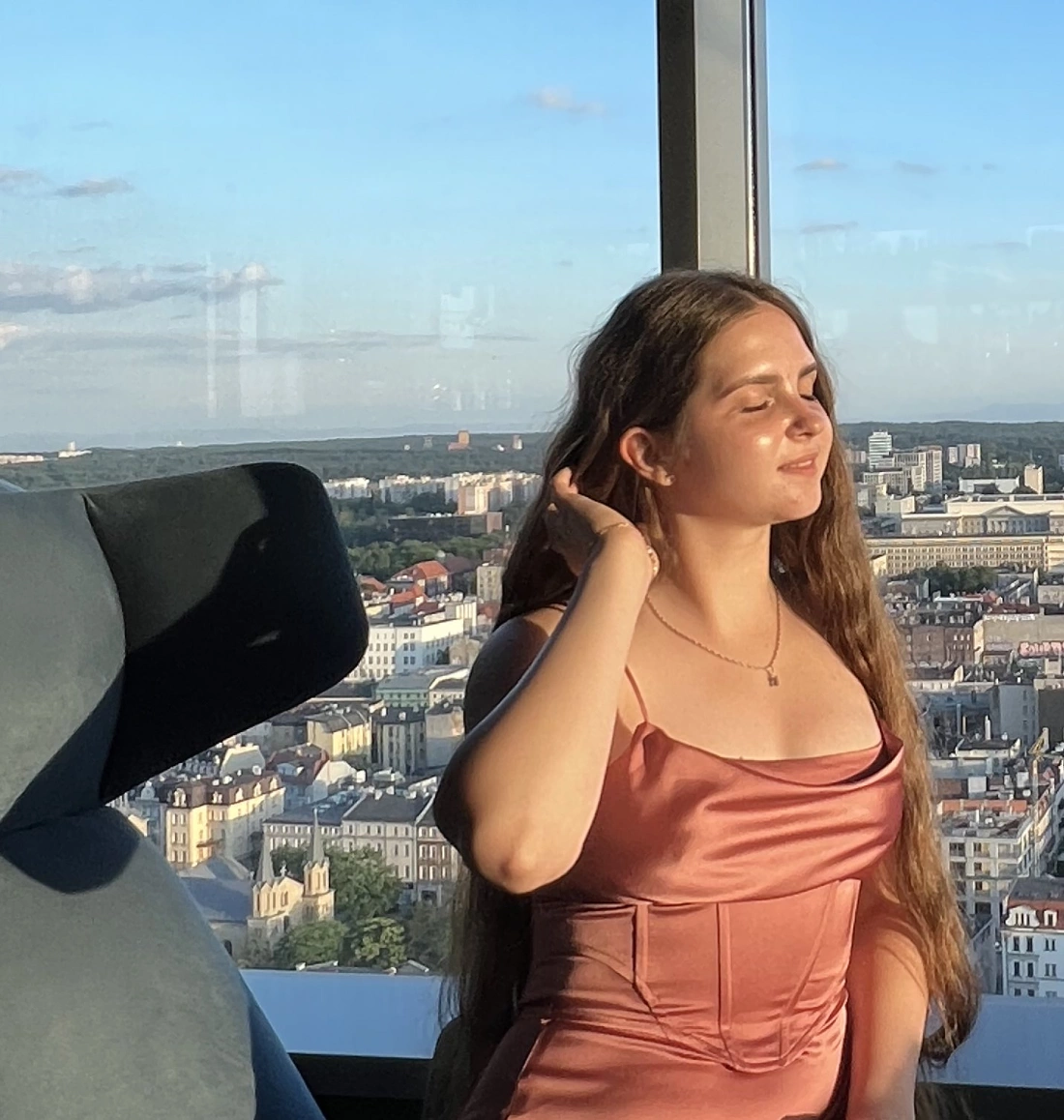 Kobieta w różowej sukience spogląda przez okno na miasto, dostrzegając panoramę w tle.