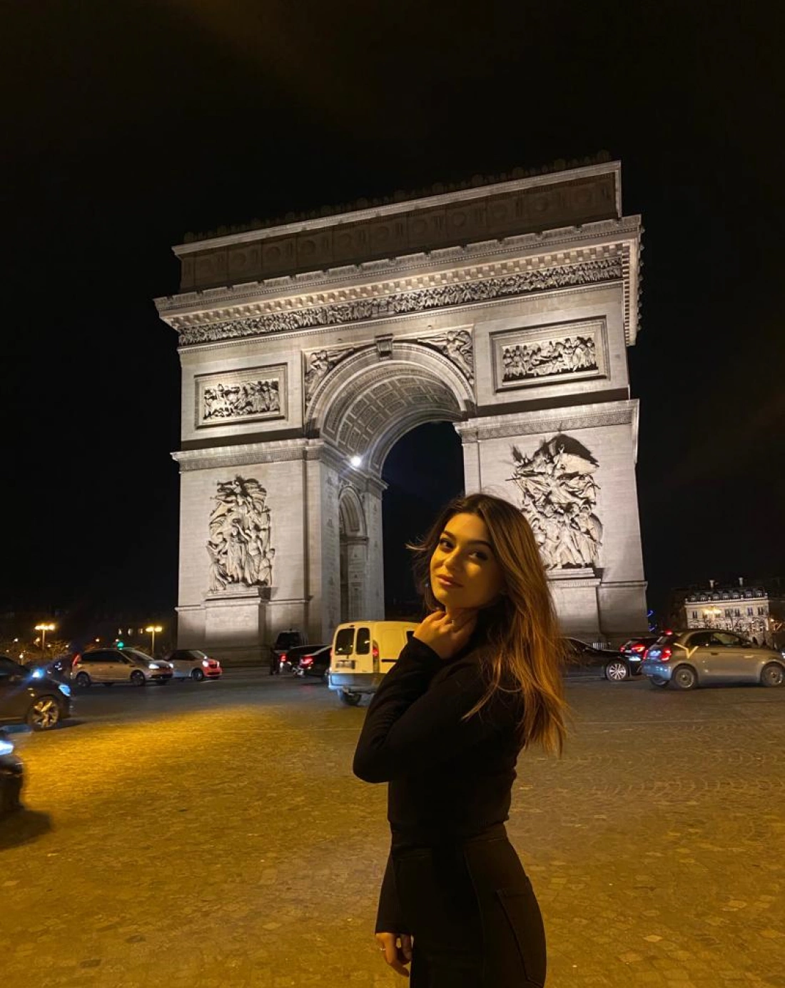 Młoda kobieta stoi przed Łukiem Triumfalnym nocą.