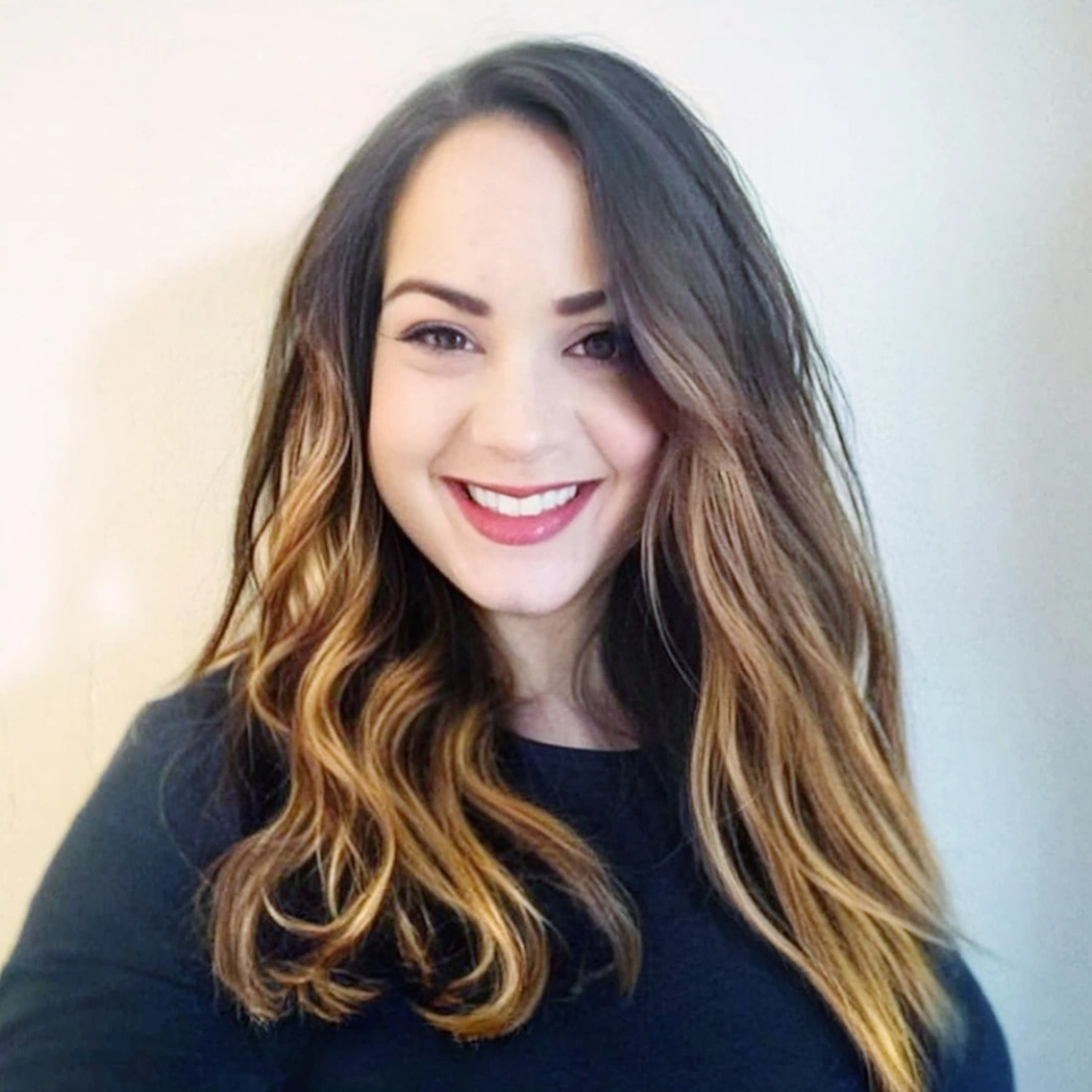 Młoda kobieta z długimi, falującymi włosami, uśmiechająca się do kamery na tle jasnej ściany.