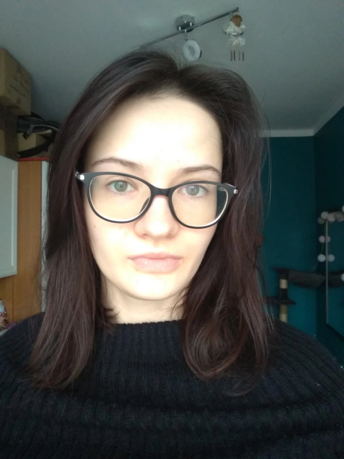 Portret młodej kobiety w czarnym swetrze i okularach, z tle w jasnych kolorach.