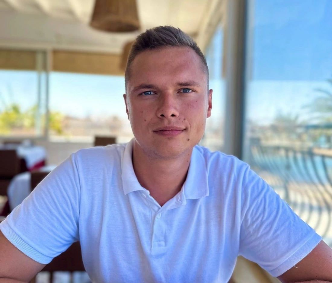 Młody mężczyzna siedzi przy stoliku w jasnej koszulce, uśmiechając się i patrząc w obiektyw, w tle widoczny jest panoramiczny widok na morze.