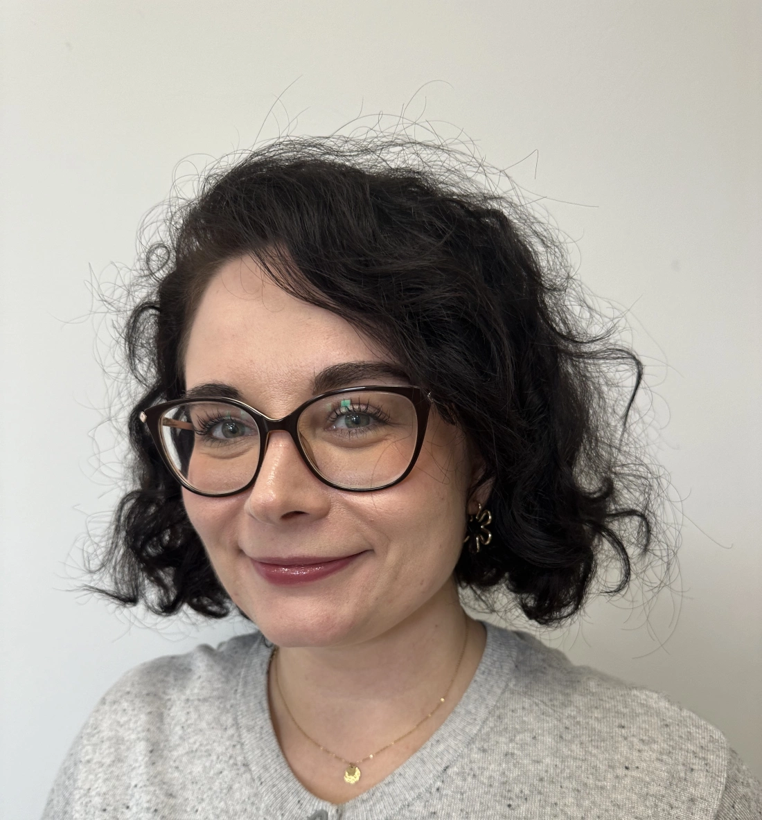 Kobieta z okularami i kręconymi włosami, uśmiechająca się, w szarej bluzie. Tło jasne.